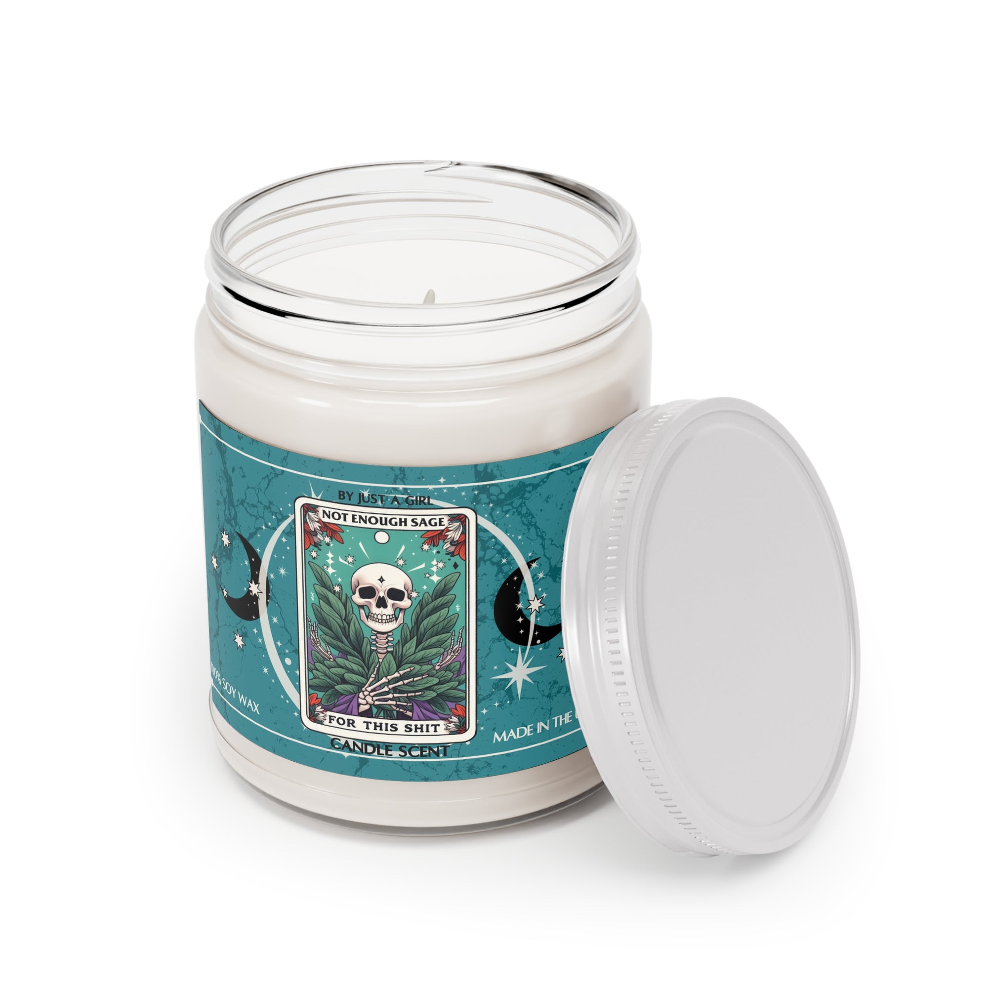 Eine weiße Kerze in einem durchsichtigen Glasgefäß mit weißem Deckel. Das Etikett ist türkisblau mit Mond- und Sternmotiven und zeigt in der Mitte eine goldgerahmte Illustration. Darauf ist ein Totenkopf zu sehen, umgeben von grünen Salbeiblättern und kleinen roten Blüten. Der Text auf dem Etikett lautet "BY JUST A GIRL".
