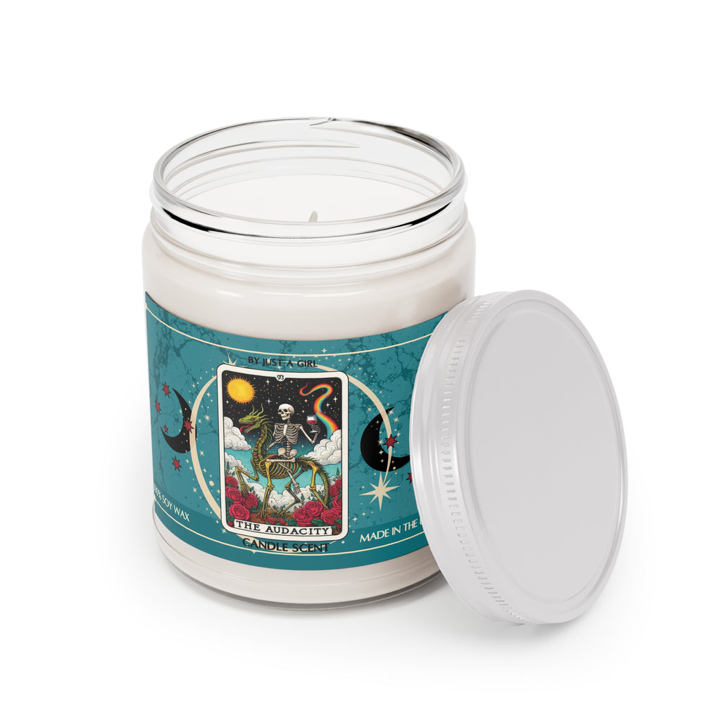 Eine weiße Kerze in einem durchsichtigen Glasgefäß mit weißem Deckel. Das Etikett ist türkisblau mit Mond- und Sternmotiven und zeigt in der Mitte eine goldgerahmte Illustration. Darauf ist ein Skelett zu sehen, das auf dem Rücken eines grünen Drachens reitet und ein rotes Weinglas in der Hand hält, umgeben von roten Rosen. Im Hintergrund ist ein sonniger Himmel mit einem Regenbogen zu sehen. Der Text auf dem Etikett lautet "BY JUST A GIRL".