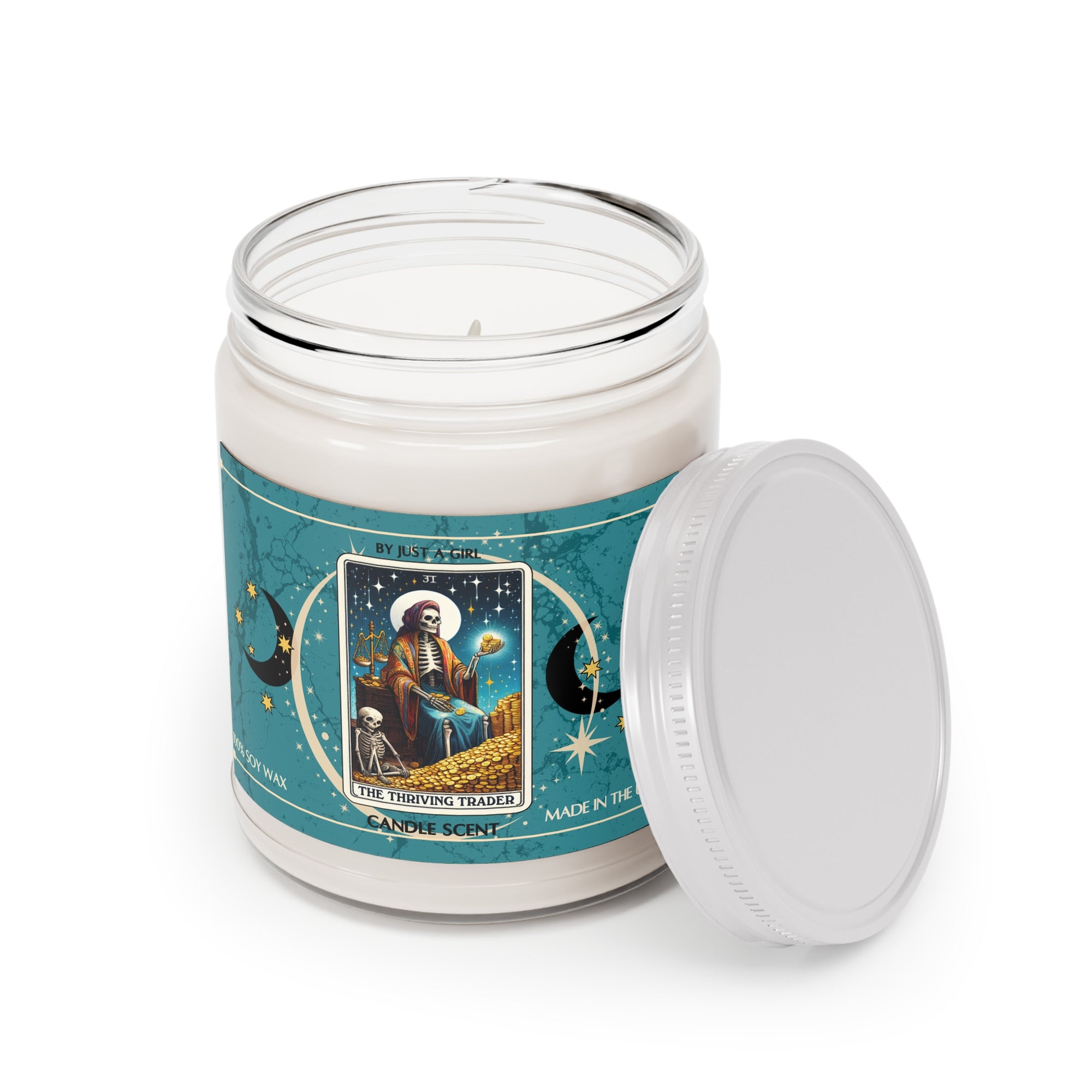 Eine weiße Kerze in einem durchsichtigen Glasgefäß mit weißem Deckel. Das Etikett ist türkisblau mit Mond- und Sternmotiven und zeigt in der Mitte eine goldgerahmte Illustration. Darauf ist ein sitzendes Skelett mit Waage und Gold in der Hand zu sehen, umgeben von einem Berg Goldmünzen und einem kleineren Skelett. Der Text auf dem Etikett lautet "BY JUST A GIRL", "31", "THE THRIVING TRADER" und "CANDLE SCENT". Unten steht "100% SOY WAX" und "MADE IN THE U.S.A.".