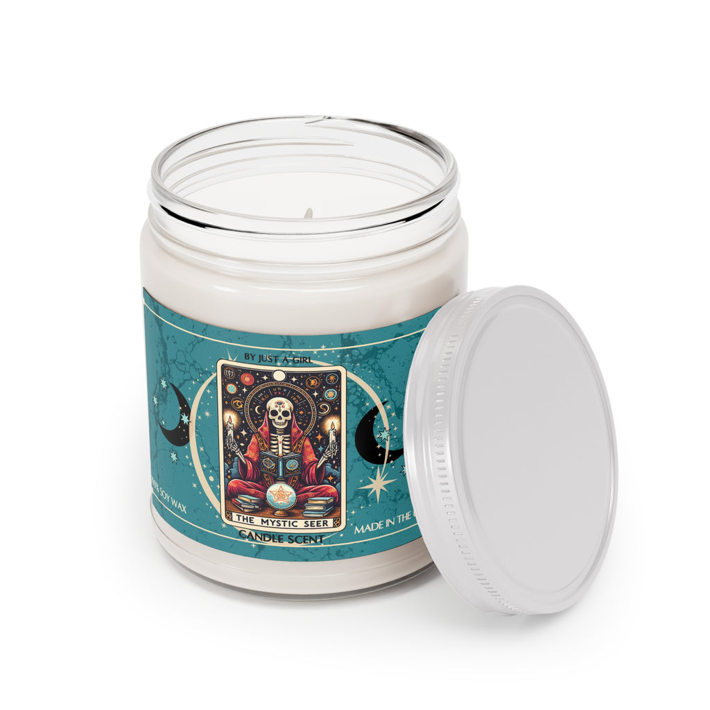 Eine weiße Kerze in einem durchsichtigen Glasgefäß mit weißem Deckel. Das Etikett ist türkisblau mit Mond- und Sternmotiven und zeigt in der Mitte eine goldgerahmte Illustration. Darauf ist ein Skelett in roter Robe zu sehen, das mit gekreuzten Beinen vor einer leuchtenden Kristallkugel sitzt und von Büchern, Kerzen und astrologischen Symbolen umgeben ist. Darüber befindet sich ein Nachthimmel mit Sternen und Planeten. Der Text auf dem Etikett lautet "BY JUST A GIRL", "THE MYSTIC SEER" und "CANDLE SCENT". 