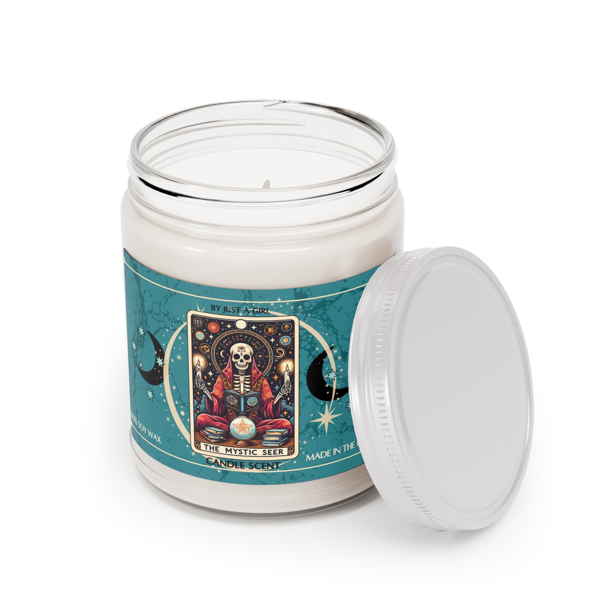 Eine weiße Kerze in einem durchsichtigen Glasgefäß mit weißem Deckel. Das Etikett ist türkisblau mit Mond- und Sternmotiven und zeigt in der Mitte eine goldgerahmte Illustration. Darauf ist ein Skelett in roter Robe zu sehen, das mit gekreuzten Beinen vor einer leuchtenden Kristallkugel sitzt und von Büchern, Kerzen und astrologischen Symbolen umgeben ist. Darüber befindet sich ein Nachthimmel mit Sternen und Planeten. Der Text auf dem Etikett lautet "BY JUST A GIRL", "THE MYSTIC SEER" und "CANDLE SCENT". 