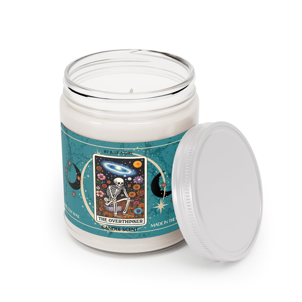 Eine weiße Kerze in einem durchsichtigen Glasgefäß mit weißem Deckel. Das Etikett ist türkisblau mit Mond- und Sternmotiven und zeigt in der Mitte eine goldgerahmte Illustration. Darauf ist ein Skelett zu sehen, das mit gekreuzten Beinen inmitten eines Feldes bunter Blumen sitzt und nachdenklich in den Nachthimmel mit Sternen, Planeten und einer Galaxie blickt. Der Text auf dem Etikett lautet "BY JUST A GIRL"