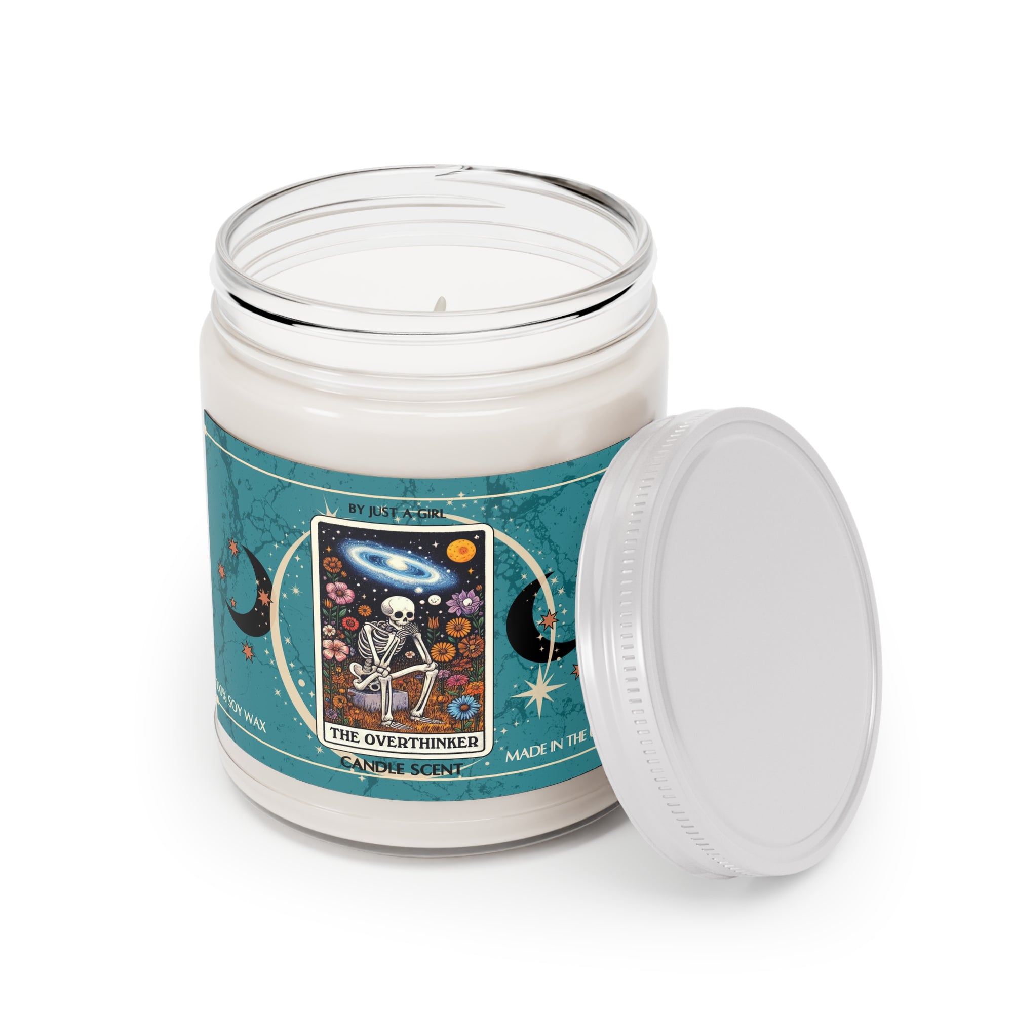 Eine weiße Kerze in einem durchsichtigen Glasgefäß mit weißem Deckel. Das Etikett ist türkisblau mit Mond- und Sternmotiven und zeigt in der Mitte eine goldgerahmte Illustration. Darauf ist ein Skelett zu sehen, das mit gekreuzten Beinen inmitten eines Feldes bunter Blumen sitzt und nachdenklich in den Nachthimmel mit Sternen, Planeten und einer Galaxie blickt. Der Text auf dem Etikett lautet "BY JUST A GIRL"