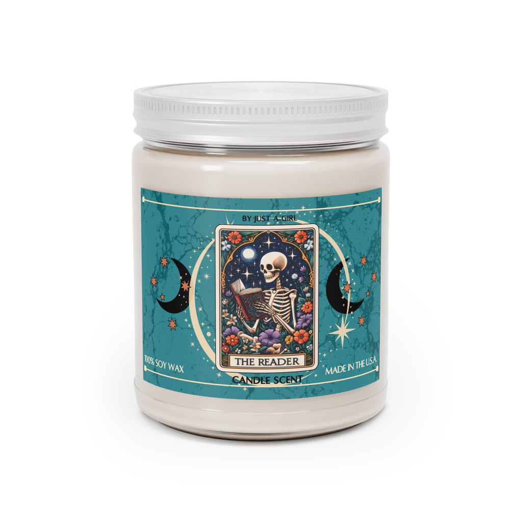 Eine weiße Kerze in einem durchsichtigen Glasgefäß mit weißem Deckel. Das Etikett ist türkisblau mit Mond- und Sternmotiven und zeigt in der Mitte eine goldgerahmte Illustration. Darauf ist ein Skelett zu sehen, das auf einem Stuhl sitzt und ein offenes Buch liest, umgeben von bunten Blumen und unter einem Mond mit Sternen. Der Text auf dem Etikett lautet "BY JUST A GIRL", 