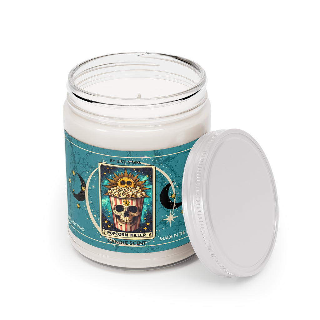 Eine weiße Kerze in einem durchsichtigen Glasgefäß mit weißem Deckel. Das Etikett ist türkisblau mit Mond- und Sternmotiven und zeigt in der Mitte eine goldgerahmte Illustration. Darauf ist ein Popcorn-Eimer abgebildet, dessen unterer Teil ein Totenkopfgesicht zeigt, gefüllt mit Popcorn, und oben eine lachende Sonne. Der Text auf dem Etikett lautet "BY JUST A GIRL".