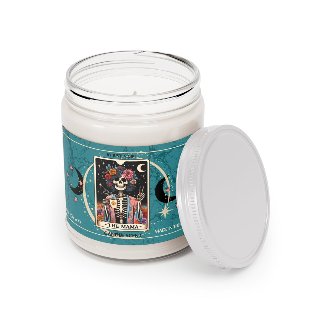 Eine weiße Kerze in einem durchsichtigen Glasgefäß mit weißem Deckel. Das Etikett ist türkisblau mit Mond- und Sternmotiven und zeigt in der Mitte eine goldgerahmte Illustration. Darauf ist ein lächelndes Skelett mit Blumen im Haar und einem bunten Blumenmuster auf der Robe zu sehen, das eine Tasse Kaffee hält und einen Peace-Zeichen mit der anderen Hand macht. Der Text auf dem Etikett lautet "BY JUST A GIRL", "THE MAMA" und "CANDLE SCENT". Unten steht "100% SOY WAX" und "MADE IN THE U.S.A.".