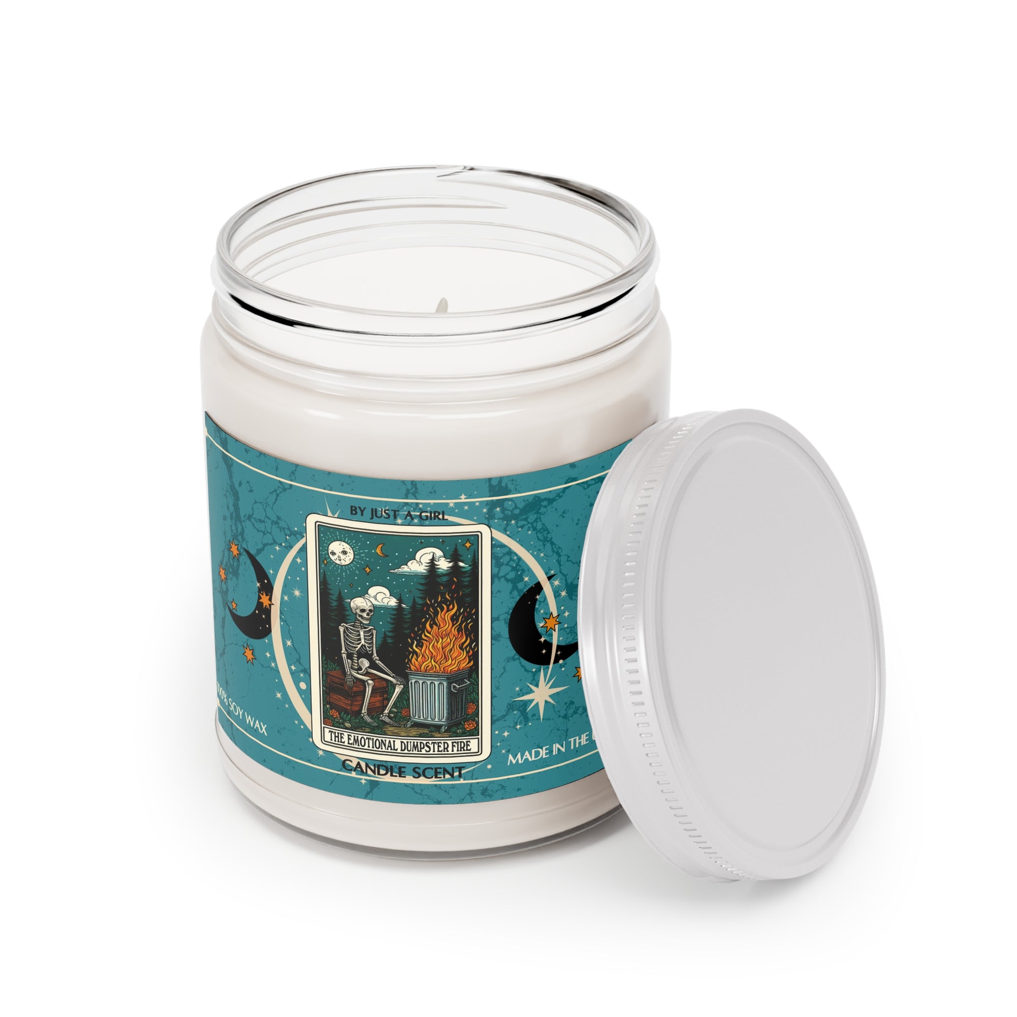 Eine weiße Kerze in einem durchsichtigen Glasgefäß mit weißem Deckel. Das Etikett ist türkisblau mit Mond- und Sternmotiven und zeigt in der Mitte eine goldgerahmte Illustration. Darauf ist ein Skelett zu sehen, das auf einer Bank vor einem brennenden Mülleimer sitzt. Über dem Skelett ist ein sternenklarer Nachthimmel mit einem Mond zu sehen. Der Text auf dem Etikett lautet "BY JUST A GIRL".