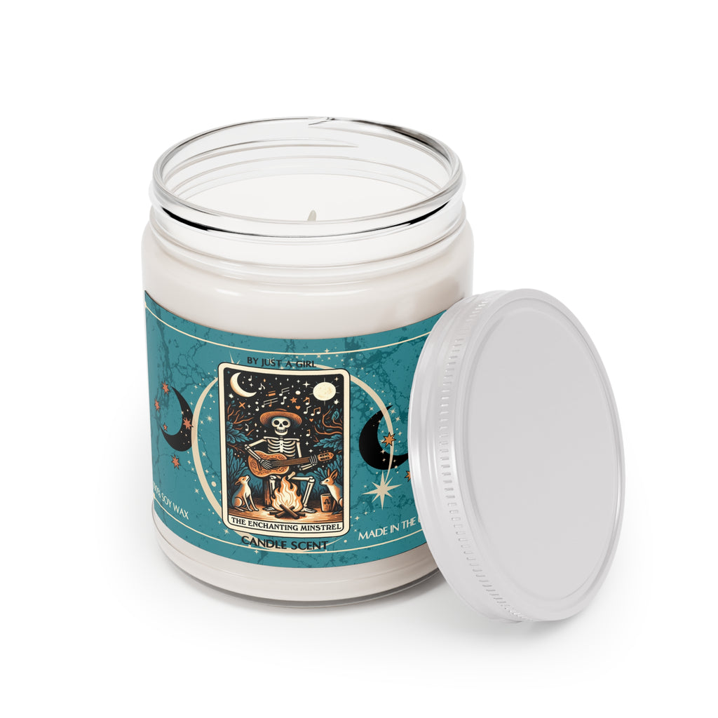 Eine weiße Kerze in einem durchsichtigen Glasgefäß mit weißem Deckel. Das Etikett ist türkisblau mit Mond- und Sternmotiven und zeigt in der Mitte eine goldgerahmte Illustration. Darauf ist ein sitzendes Skelett mit Hut zu sehen, das Gitarre spielt, während es an einem Lagerfeuer sitzt. Zwei kleine Hunde oder Füchse sitzen daneben. Über dem Skelett ist ein sternenklarer Nachthimmel mit einem Vollmond und Noten zu sehen.