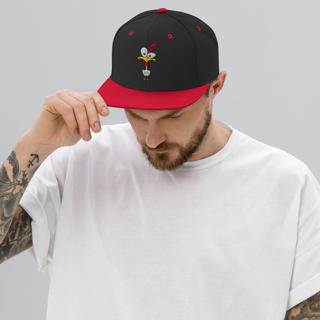 Eine klassische, zweifarbige Snapback-Cap mit flachem Schirm. Auf der Vorderseite ist ein gesticktes Cartoon-Motiv eines Huhns zu sehen, das einen hellen Kopf, große Augen, einen farbigen Kamm und einen Schnabel sowie eine kleine stilisierte Geste am Hals hat. Der obere Teil der Kappe ist dunkel, während der Schirm und der obere Knopf eine leuchtende Kontrastfarbe aufweisen.