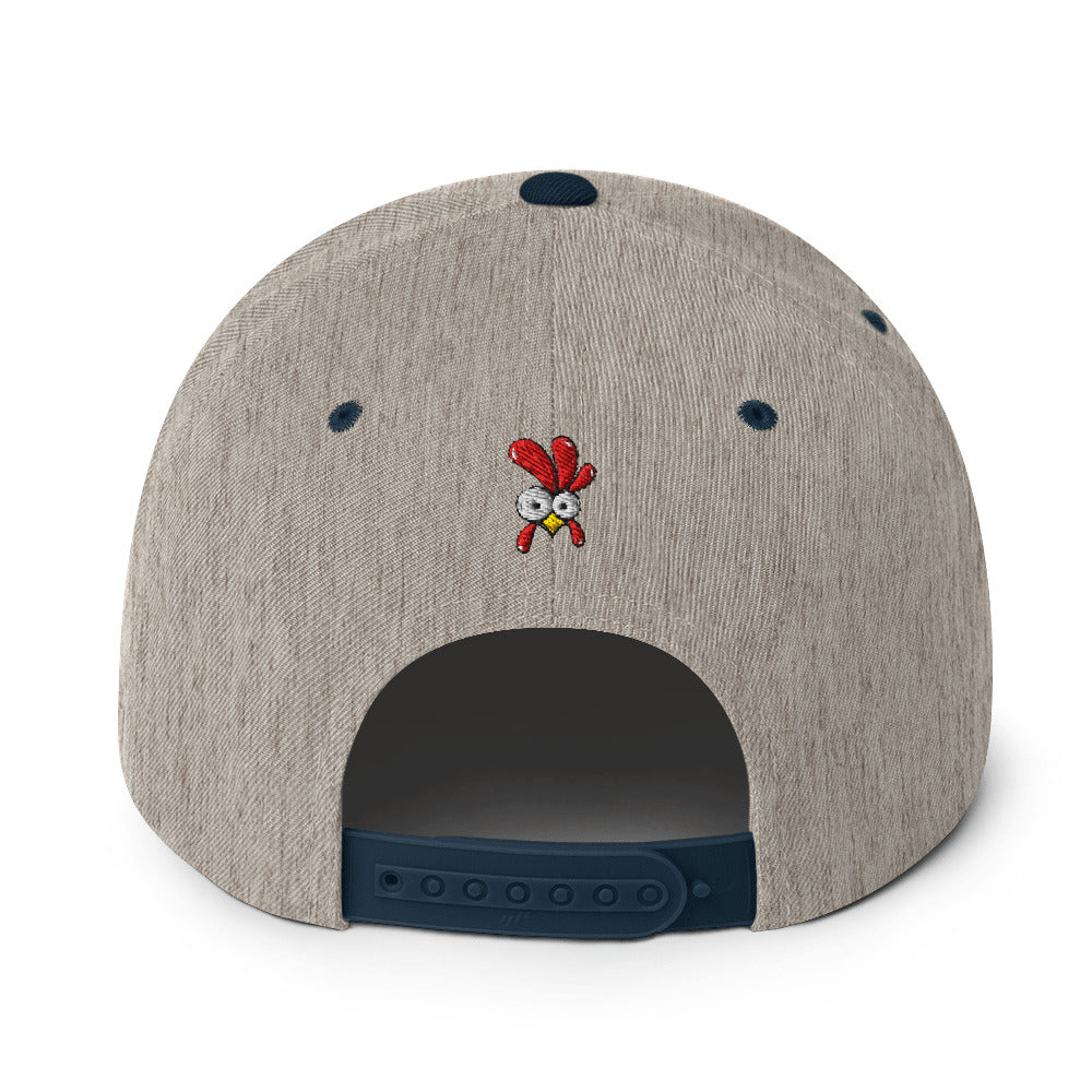 Eine klassische, zweifarbige Snapback-Cap mit flachem Schirm. Auf der Vorderseite ist ein gesticktes Cartoon-Motiv eines Huhns zu sehen, das einen hellen Kopf, große Augen, einen farbigen Kamm und einen Schnabel sowie eine kleine stilisierte Geste am Hals hat. Der obere Teil der Kappe ist dunkel, während der Schirm und der obere Knopf eine leuchtende Kontrastfarbe aufweisen.