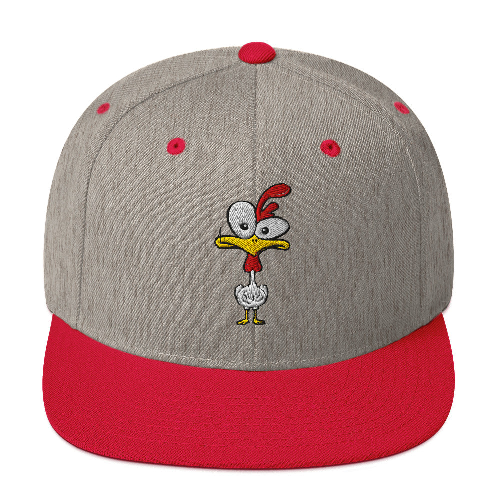 Eine klassische, zweifarbige Snapback-Cap mit flachem Schirm. Auf der Vorderseite ist ein gesticktes Cartoon-Motiv eines Huhns zu sehen, das einen hellen Kopf, große Augen, einen farbigen Kamm und einen Schnabel sowie eine kleine stilisierte Geste am Hals hat. Der obere Teil der Kappe ist dunkel, während der Schirm und der obere Knopf eine leuchtende Kontrastfarbe aufweisen.
