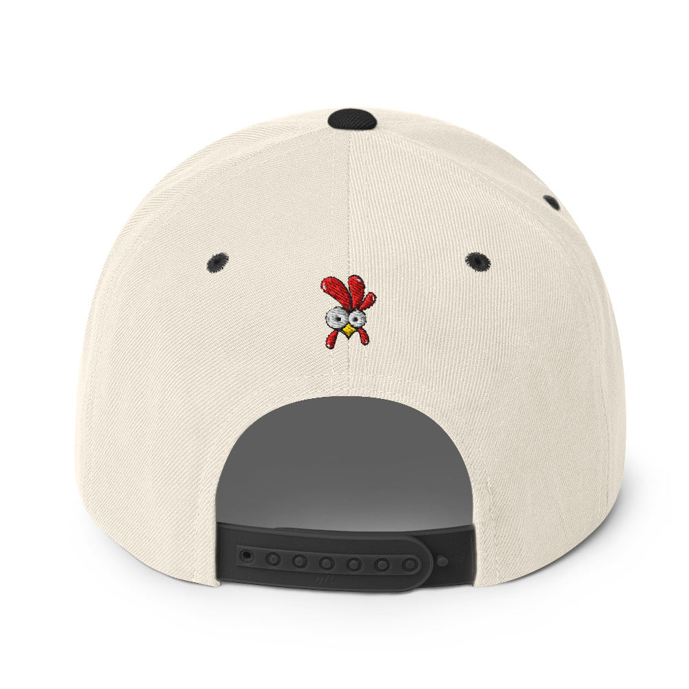 Eine klassische, zweifarbige Snapback-Cap mit flachem Schirm. Auf der Vorderseite ist ein gesticktes Cartoon-Motiv eines Huhns zu sehen, das einen hellen Kopf, große Augen, einen farbigen Kamm und einen Schnabel sowie eine kleine stilisierte Geste am Hals hat. Der obere Teil der Kappe ist dunkel, während der Schirm und der obere Knopf eine leuchtende Kontrastfarbe aufweisen.