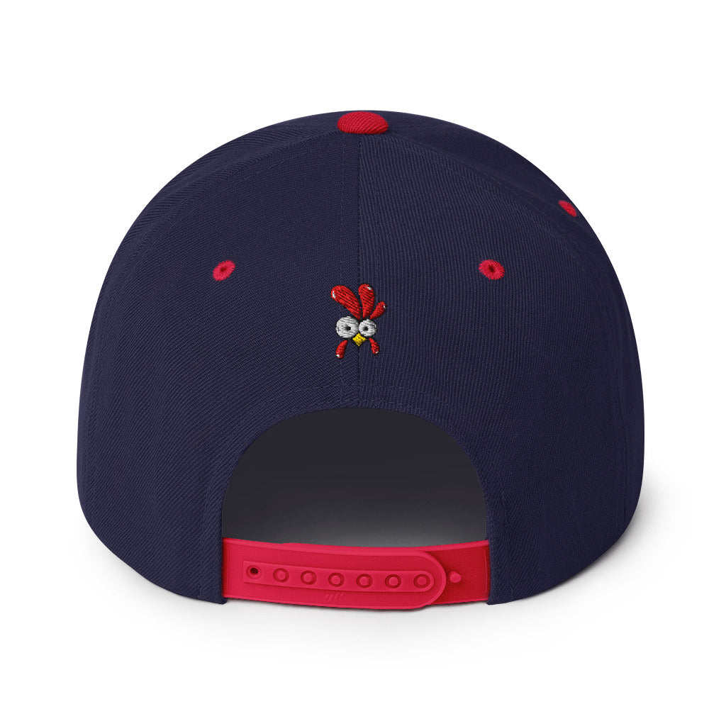Eine klassische, zweifarbige Snapback-Cap mit flachem Schirm. Auf der Vorderseite ist ein gesticktes Cartoon-Motiv eines Huhns zu sehen, das einen hellen Kopf, große Augen, einen farbigen Kamm und einen Schnabel sowie eine kleine stilisierte Geste am Hals hat. Der obere Teil der Kappe ist dunkel, während der Schirm und der obere Knopf eine leuchtende Kontrastfarbe aufweisen.