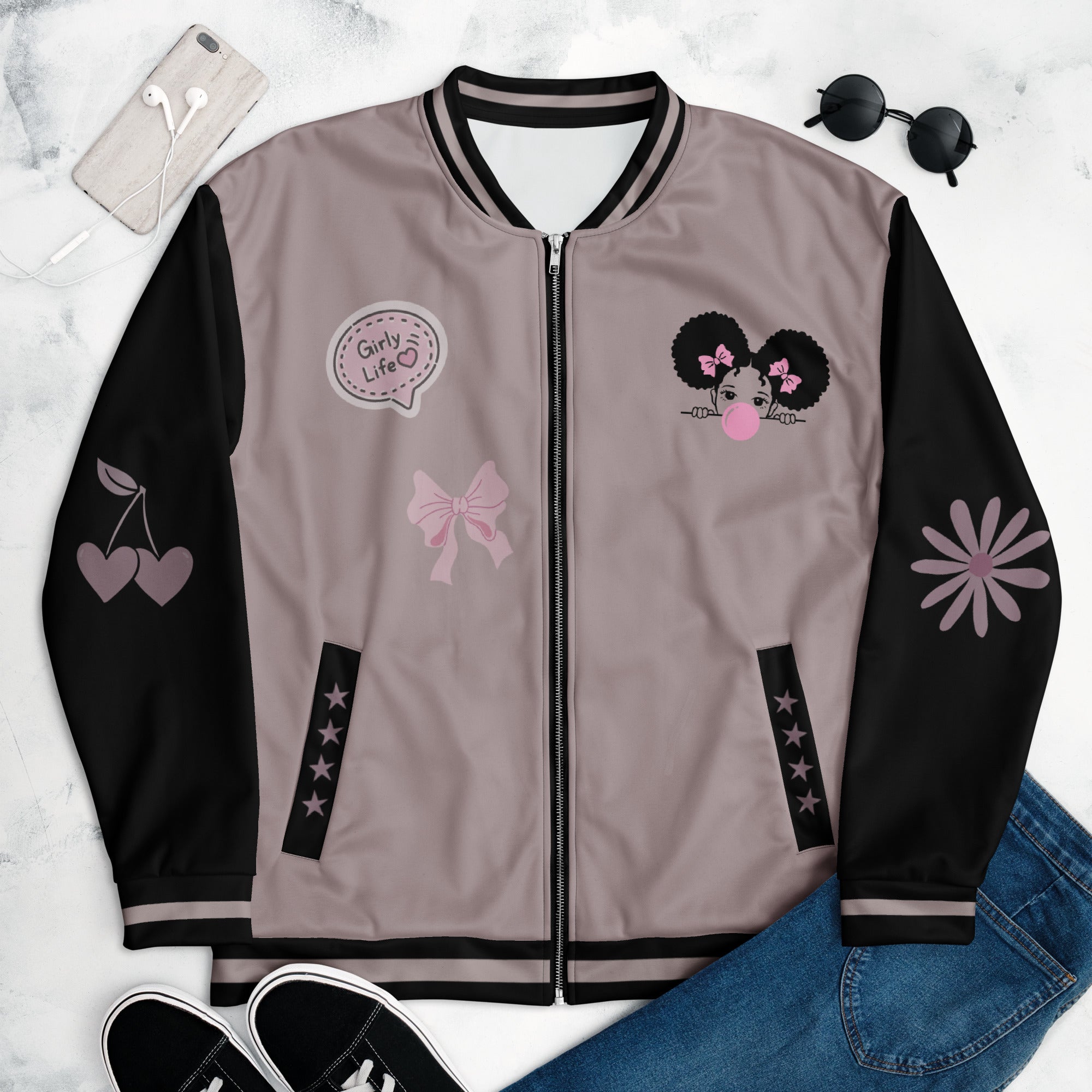 Eine Taupe farbene College Bomberjacke  mit schwarzen Ärmeln, Kragen und Saumbündchen jweils mit taupefarbenen streifen versehen. Auf der linken Brustseite ist eine Cartoon-Figur eines Mädchens mit dunklen Afro-Haaren, pinken Haarschleifen und einer großen rosa Kaugummi-Blase am Mund zu sehen. Die Jacke hat einen schwarzen Reißverschluss und schwarze Tascheneingriffe mit Sternchen an der Vorderseite. Mockup Jacke liegend vorderseite präsentiert mit Jeans, Schuhe & Sonnenbrille. By Just a Girl. 