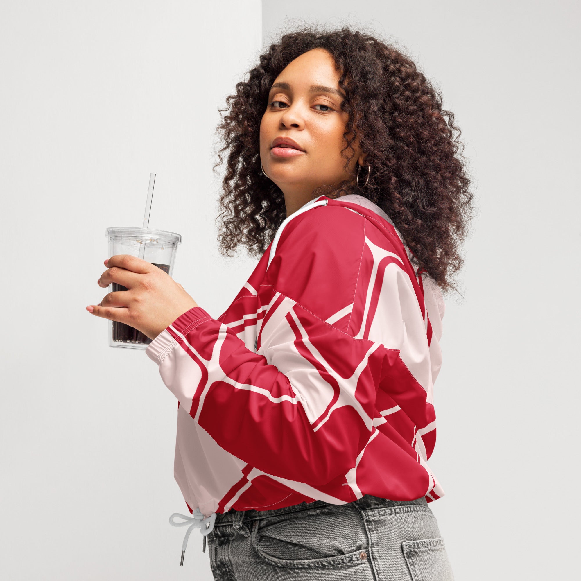 Ein Damen Cropped Windbreaker in Rot und hellem Rosé mit einem abstrakten geometrischen Muster aus miteinander verbundenen, abgerundeten Rechtecken und Linien. Die Jacke hat eine Kapuze, einen halben Reißverschluss, weiße Kordeln und einen verstellbaren Saum. Mockup woman seitliche Ansicht rechts. By Just a Girl
