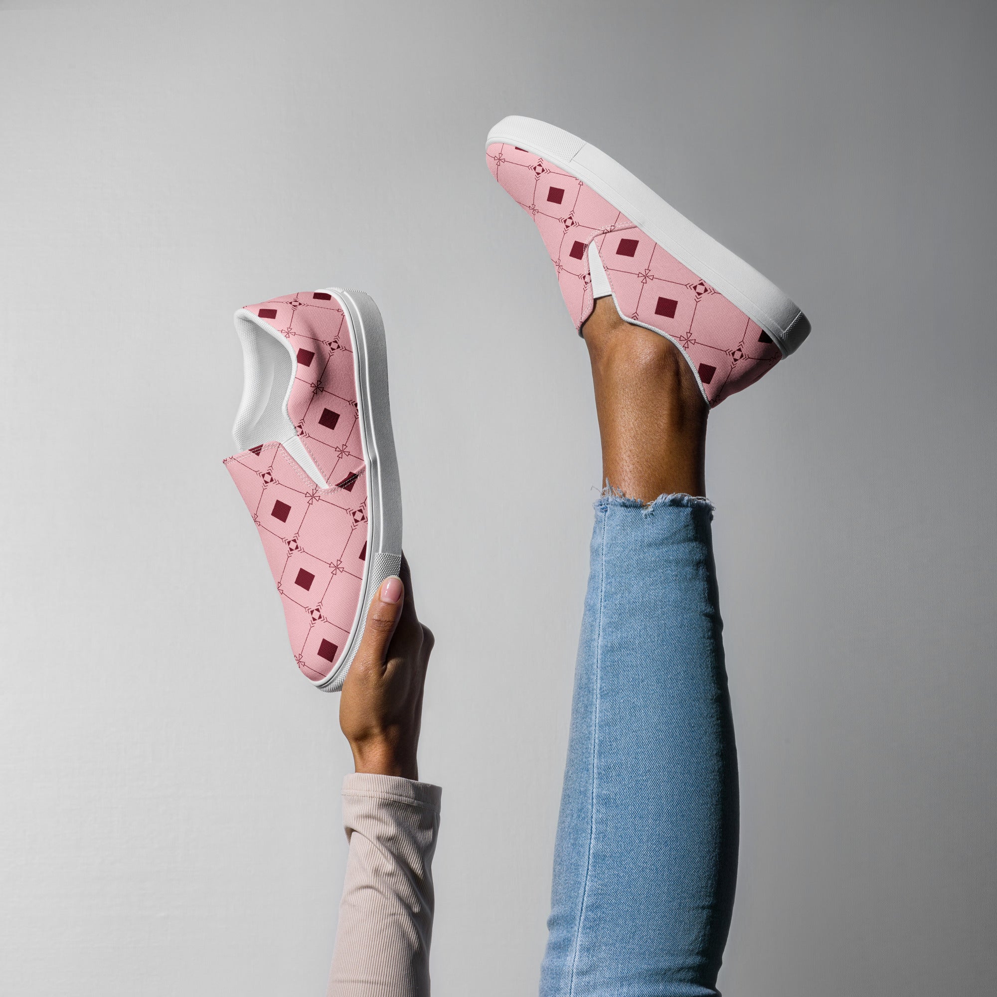 Ein weibliches Model zeigt tragend ein Bein in die Luft gestreckt und mit der anderen Hand den 2. Schuh zeigend mit einem geometrischen Muster aus dunklen roten Quadraten und Linien auf einem hellrosa Hintergrund.  Designed, by Just a Girl.
