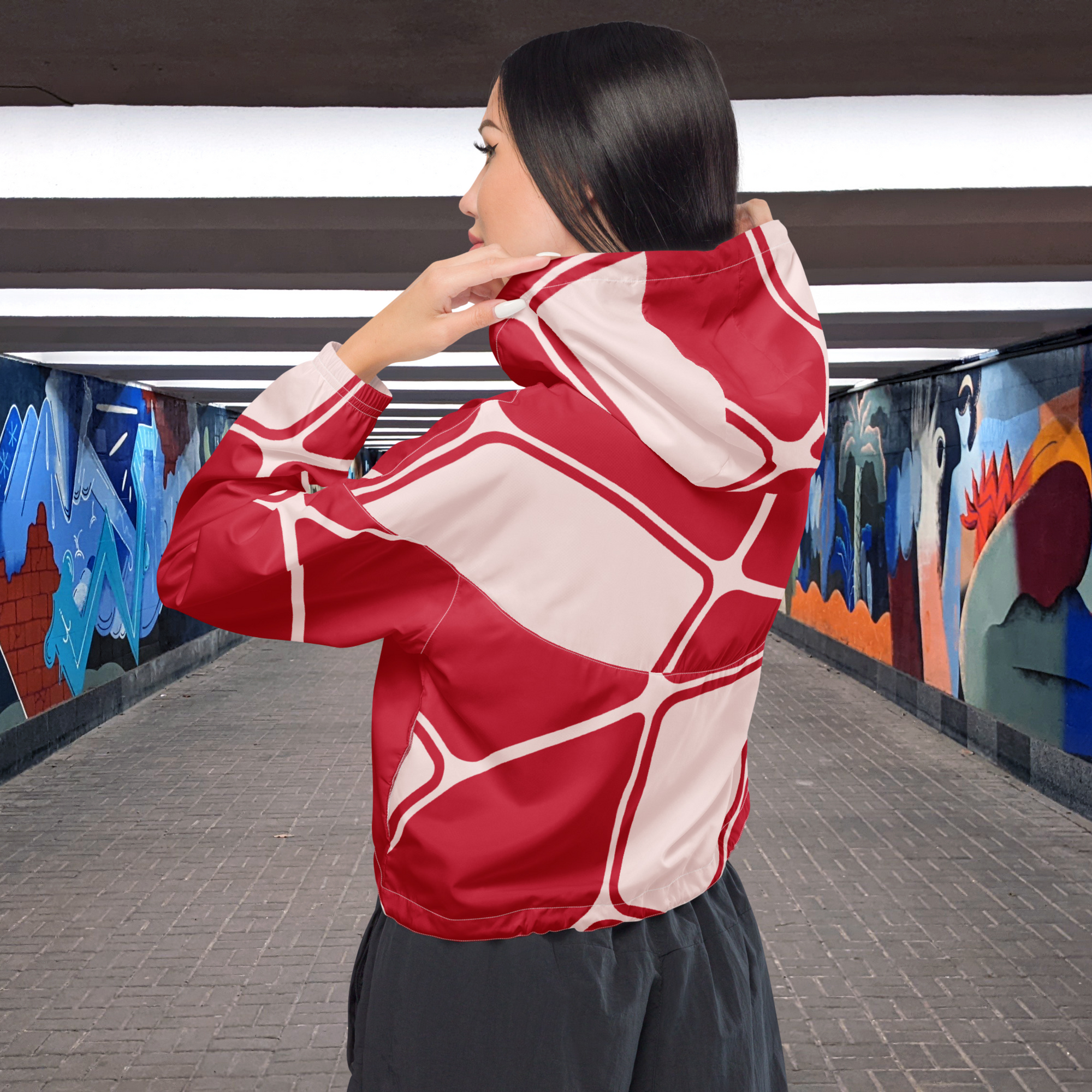 Ein Damen Cropped Windbreaker in Rot und hellem Rosé mit einem abstrakten geometrischen Muster aus miteinander verbundenen, abgerundeten Rechtecken und Linien. Die Jacke hat eine Kapuze, einen halben Reißverschluss, weiße Kordeln und einen verstellbaren Saum. Mockup Girl Rückenansicht. By Just a Girl.