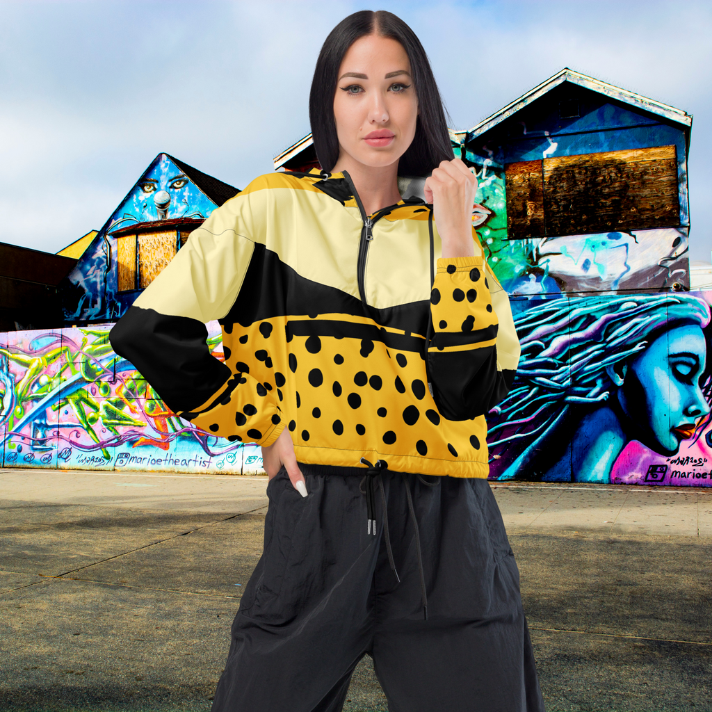 Ein Damen Cropped Windbreaker in Gelb, Schwarz und einem pastell gelb, mit einem auffälligen schwarzen Punktemuster auf der unteren gelben Hälfte. Die Jacke hat eine Kapuze, einen halben Reißverschluss, schwarze Kordeln und einen verstellbaren Saum. gezeigt von einem weiblichen mockup in der vorder ansicht im freien. By Just a Girl.