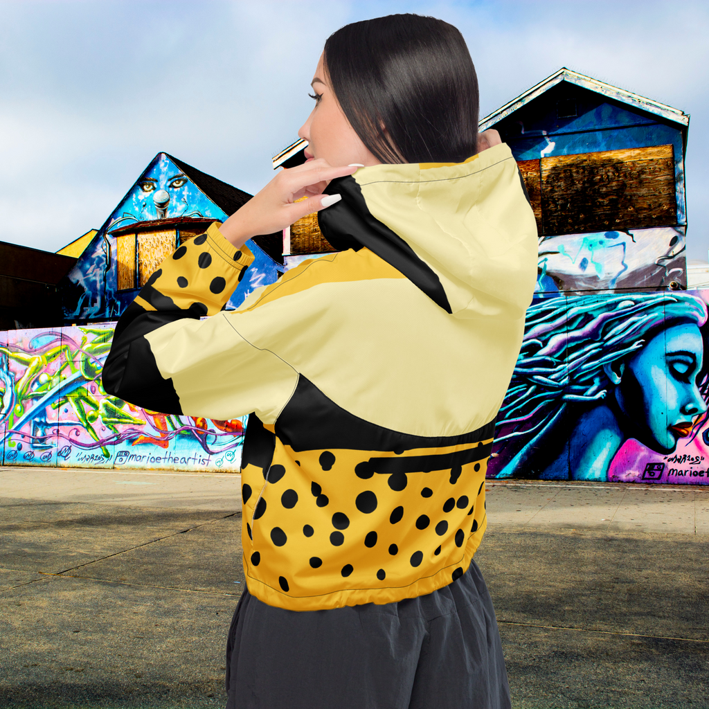 Ein Damen Cropped Windbreaker in Gelb, Schwarz und einem pastell gelb, mit einem auffälligen schwarzen Punktemuster auf der unteren gelben Hälfte. Die Jacke hat eine Kapuze, einen halben Reißverschluss, schwarze Kordeln und einen verstellbaren Saum. gezeigt von einem weiblichen mockup in der rückansicht im freien. By Just a Girl.
