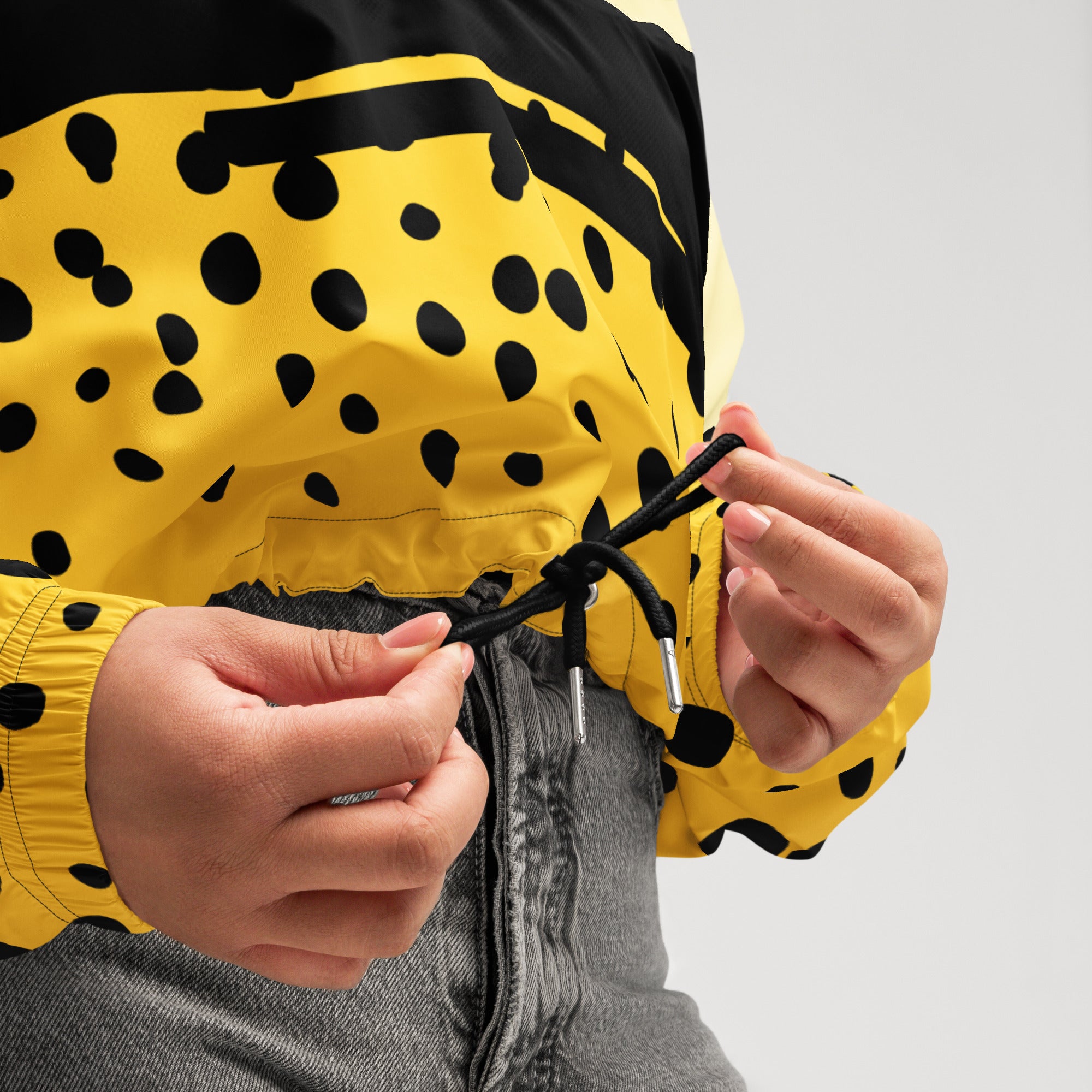 Ein Damen Cropped Windbreaker in Gelb, Schwarz und einem pastell gelb, mit einem auffälligen schwarzen Punktemuster auf der unteren gelben Hälfte. Die Jacke hat eine Kapuze, einen halben Reißverschluss, schwarze Kordeln und einen verstellbaren Saum.  By Just a Girl.