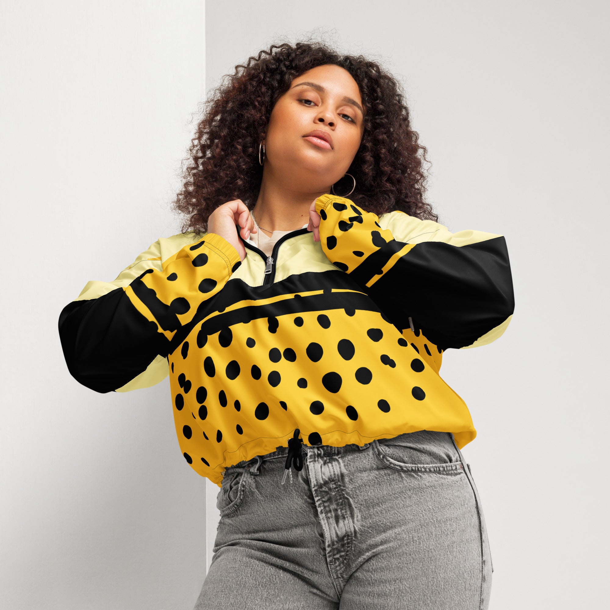 Ein Damen Cropped Windbreaker in Gelb, Schwarz und einem pastell gelb, mit einem auffälligen schwarzen Punktemuster auf der unteren gelben Hälfte. Die Jacke hat eine Kapuze, einen halben Reißverschluss, schwarze Kordeln und einen verstellbaren Saum. gezeigt von einer kräftigeren weiblichen mockup in der vorder ansicht. By Just a Girl.