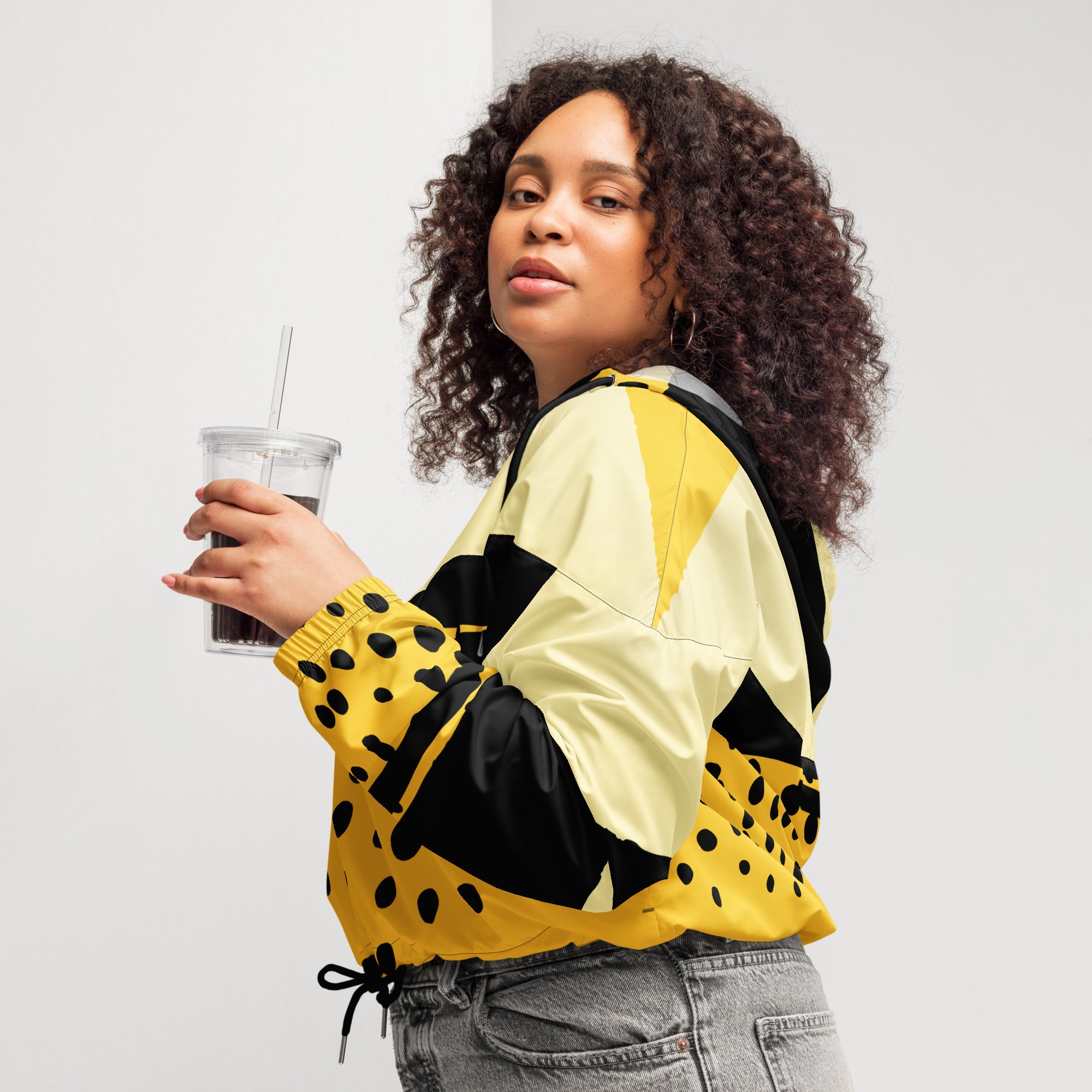 Ein Damen Cropped Windbreaker in Gelb, Schwarz und einem pastell gelb, mit einem auffälligen schwarzen Punktemuster auf der unteren gelben Hälfte. Die Jacke hat eine Kapuze, einen halben Reißverschluss, schwarze Kordeln und einen verstellbaren Saum. gezeigt von einer kräftigeren weiblichen mockup in der linken seiten ansicht. By Just a Girl.