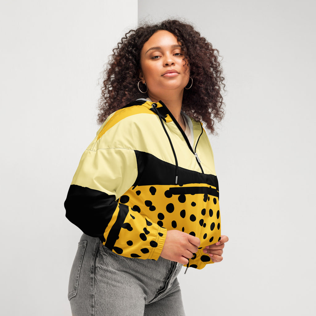 Ein Damen Cropped Windbreaker in Gelb, Schwarz und einem pastell gelb, mit einem auffälligen schwarzen Punktemuster auf der unteren gelben Hälfte. Die Jacke hat eine Kapuze, einen halben Reißverschluss, schwarze Kordeln und einen verstellbaren Saum. gezeigt von einer kräftigeren weiblichen mockup in der rechten seitenansicht im freien. By Just a Girl.