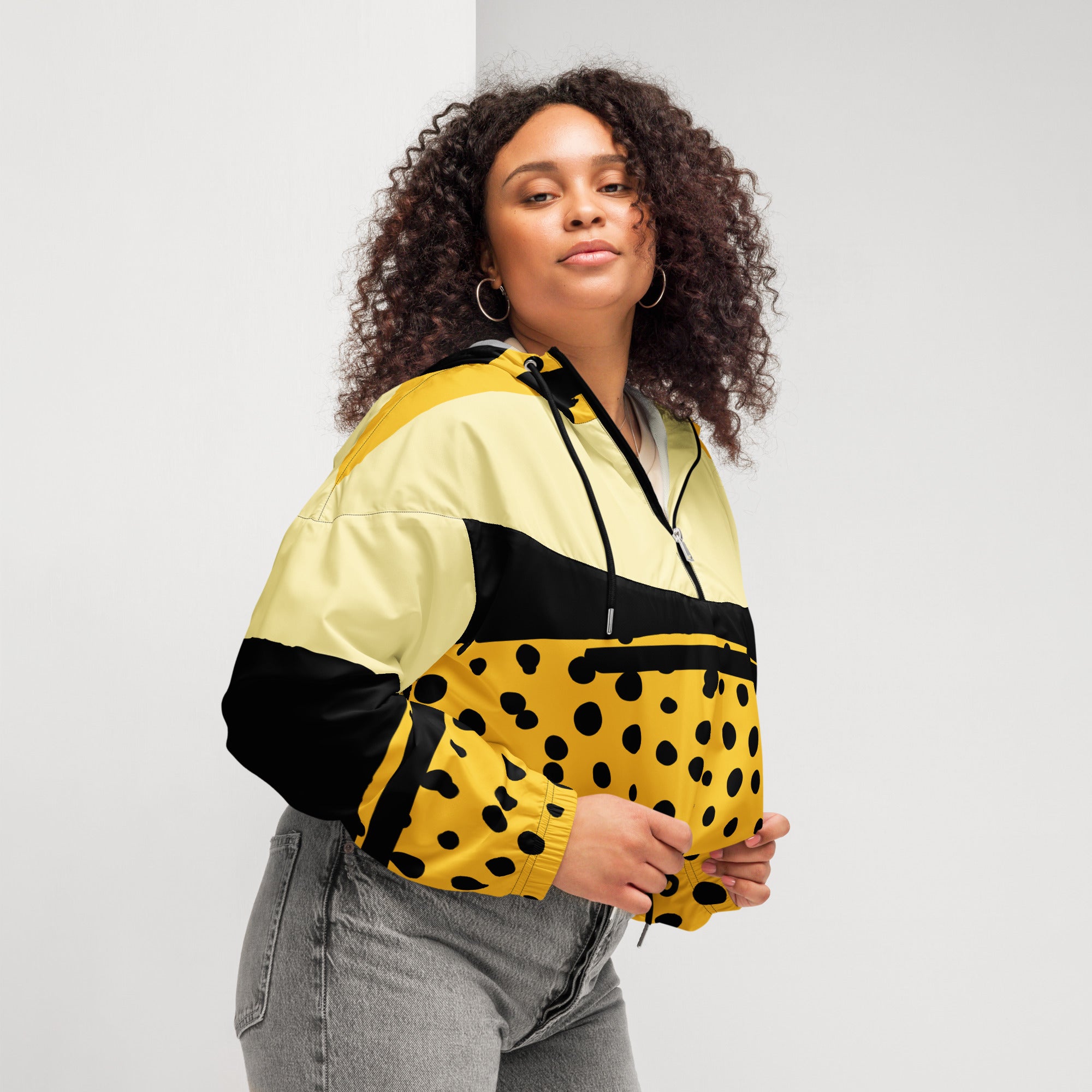 Ein Damen Cropped Windbreaker in Gelb, Schwarz und einem pastell gelb, mit einem auffälligen schwarzen Punktemuster auf der unteren gelben Hälfte. Die Jacke hat eine Kapuze, einen halben Reißverschluss, schwarze Kordeln und einen verstellbaren Saum. gezeigt von einer kräftigeren weiblichen mockup in der rechten seitenansicht im freien. By Just a Girl.