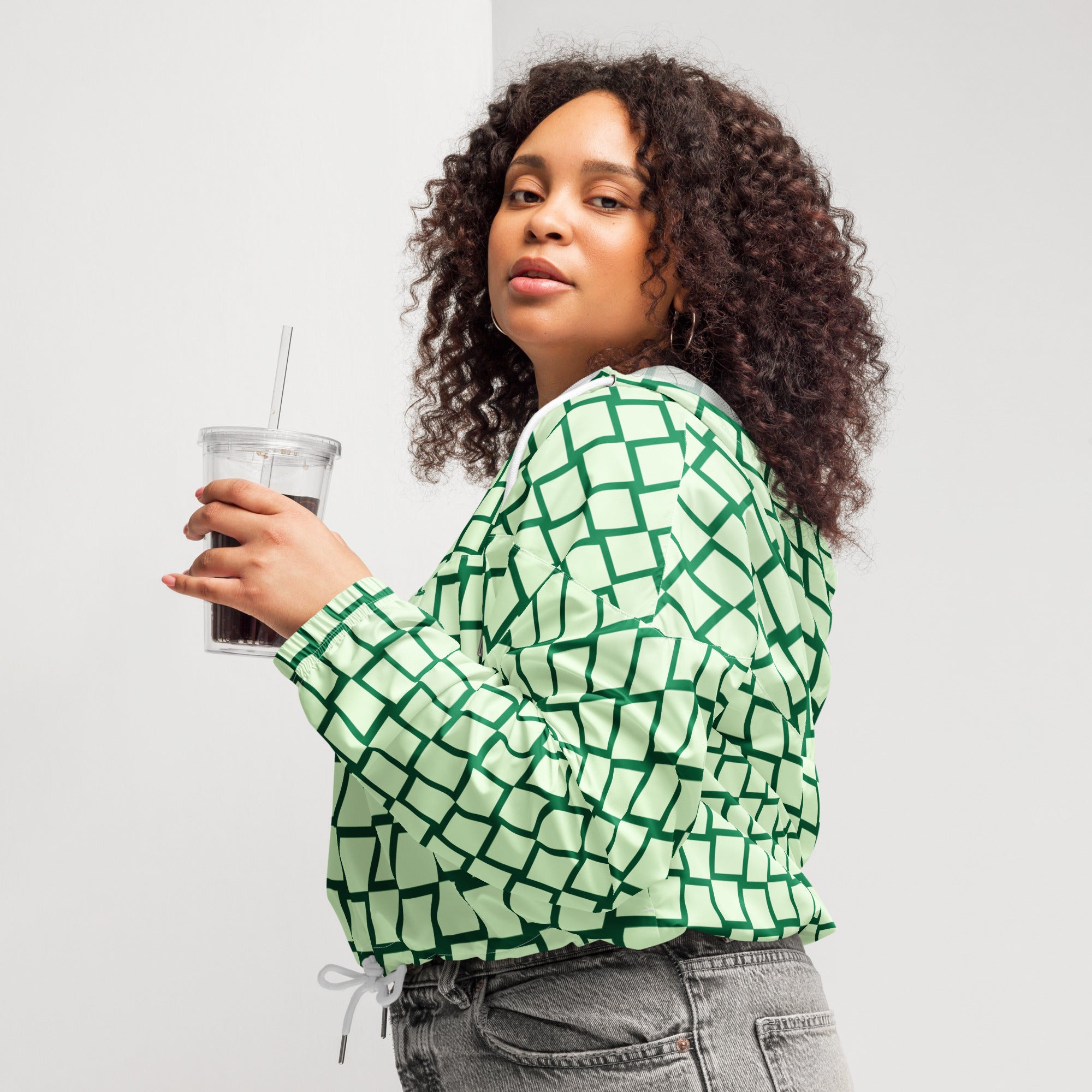 Ein Damen Cropped Windbreaker mit einem Schachbrettmuster in hellem Mintgrün und dunklem Grün. Die Jacke hat eine Kapuze, einen halben Reißverschluss mit weißen Kordeln und einen verstellbaren Saum. Mockup woman seitliche Ansicht links. By Just a Girl.
