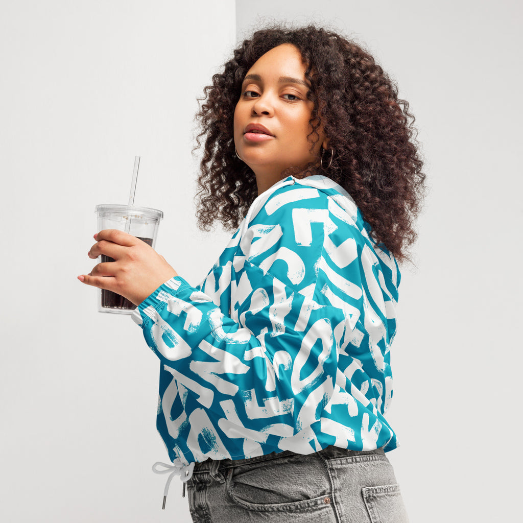 Ein Damen Cropped Windbreaker in Türkis mit einem Muster aus großen, weißen und unregelmäßig geformten Buchstaben. Die Jacke hat eine Kapuze, einen halben Reißverschluss, weiße Kordeln und einen verstellbaren Saum. Die Jacke ist flach ausgebreitet. Angezogen an einer kräftigeren Frau seitlich by Just a Girl.