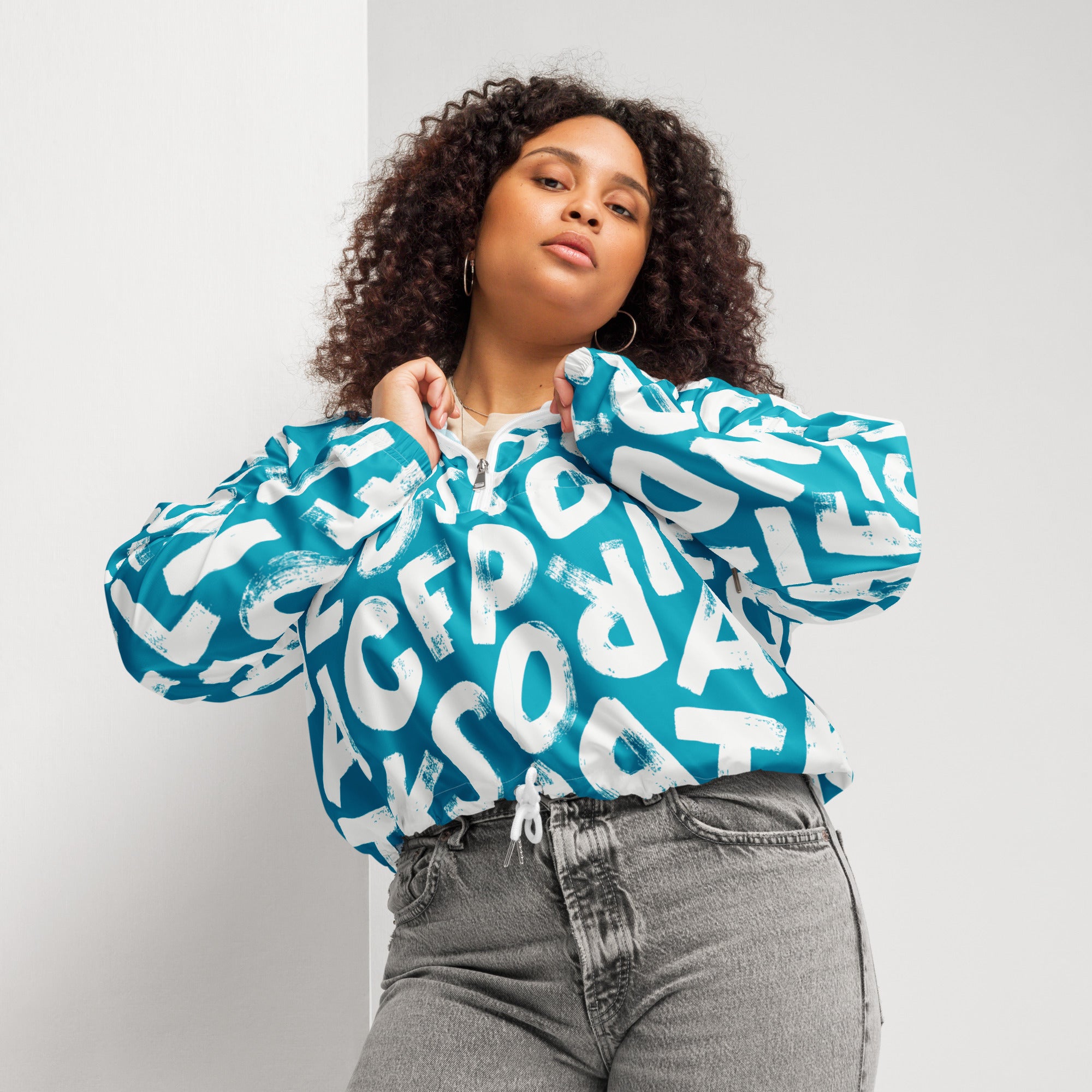 Ein Damen Cropped Windbreaker in Türkis mit einem Muster aus großen, weißen und unregelmäßig geformten Buchstaben. Die Jacke hat eine Kapuze, einen halben Reißverschluss, weiße Kordeln und einen verstellbaren Saum. Die Jacke ist flach ausgebreitet. Angezogen an einer kräftigeren Frau von Vorne by Just a Girl.
