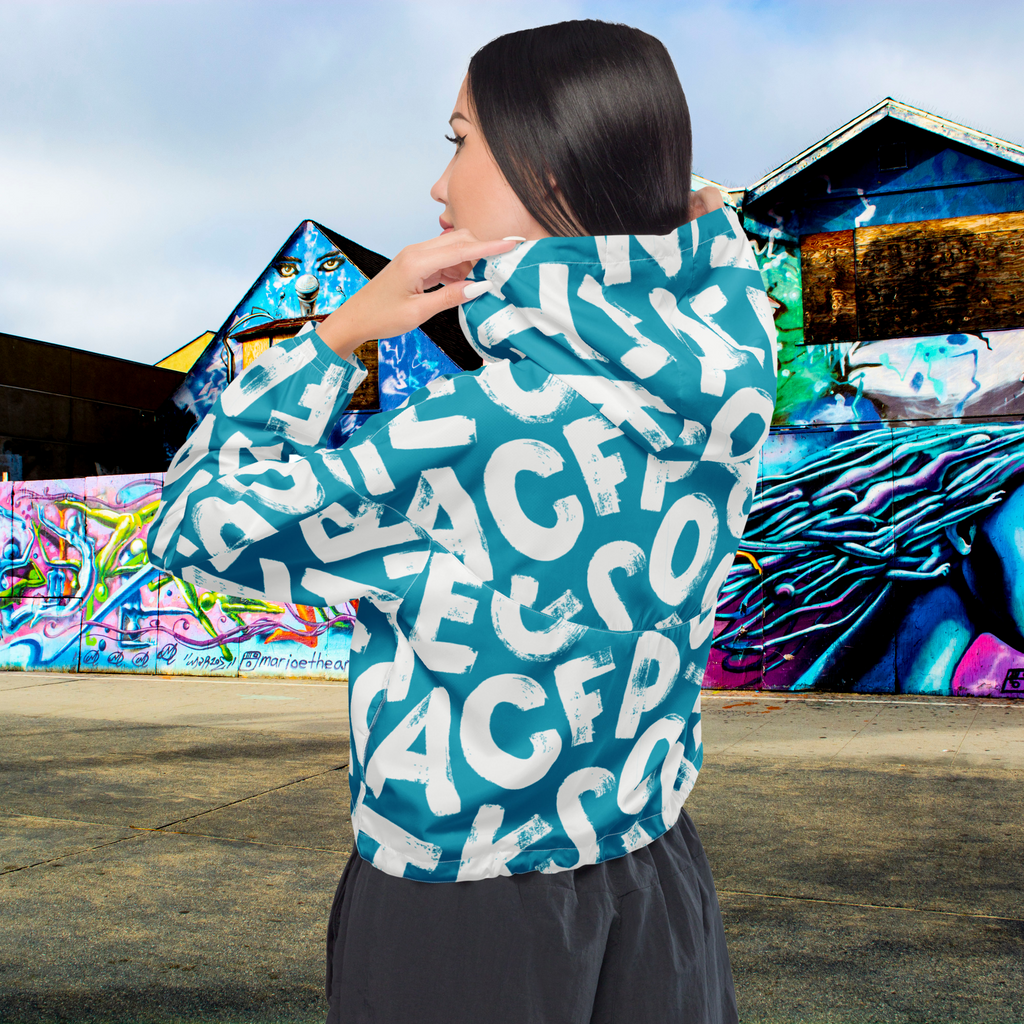Ein Damen Cropped Windbreaker in Türkis mit einem Muster aus großen, weißen und unregelmäßig geformten Buchstaben. Die Jacke hat eine Kapuze, einen halben Reißverschluss, weiße Kordeln und einen verstellbaren Saum. Die Jacke ist flach ausgebreitet. Angezogen an einer Frau sichtbar von hinten  by Just a Girl.