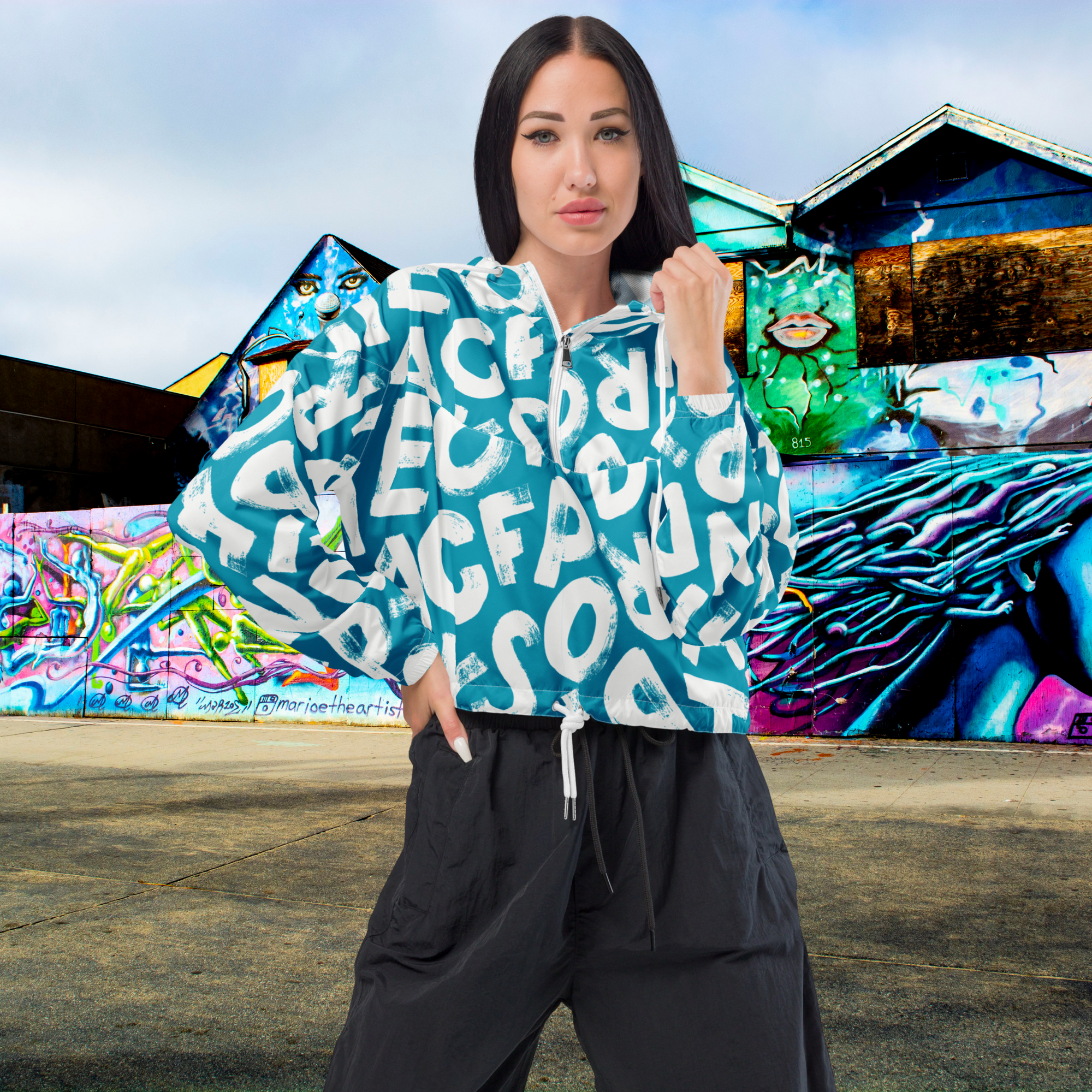 Ein Damen Cropped Windbreaker in Türkis mit einem Muster aus großen, weißen und unregelmäßig geformten Buchstaben. Die Jacke hat eine Kapuze, einen halben Reißverschluss, weiße Kordeln und einen verstellbaren Saum. Die Jacke ist flach ausgebreitet. Angezogen an einer Frau. by Just a Girl.