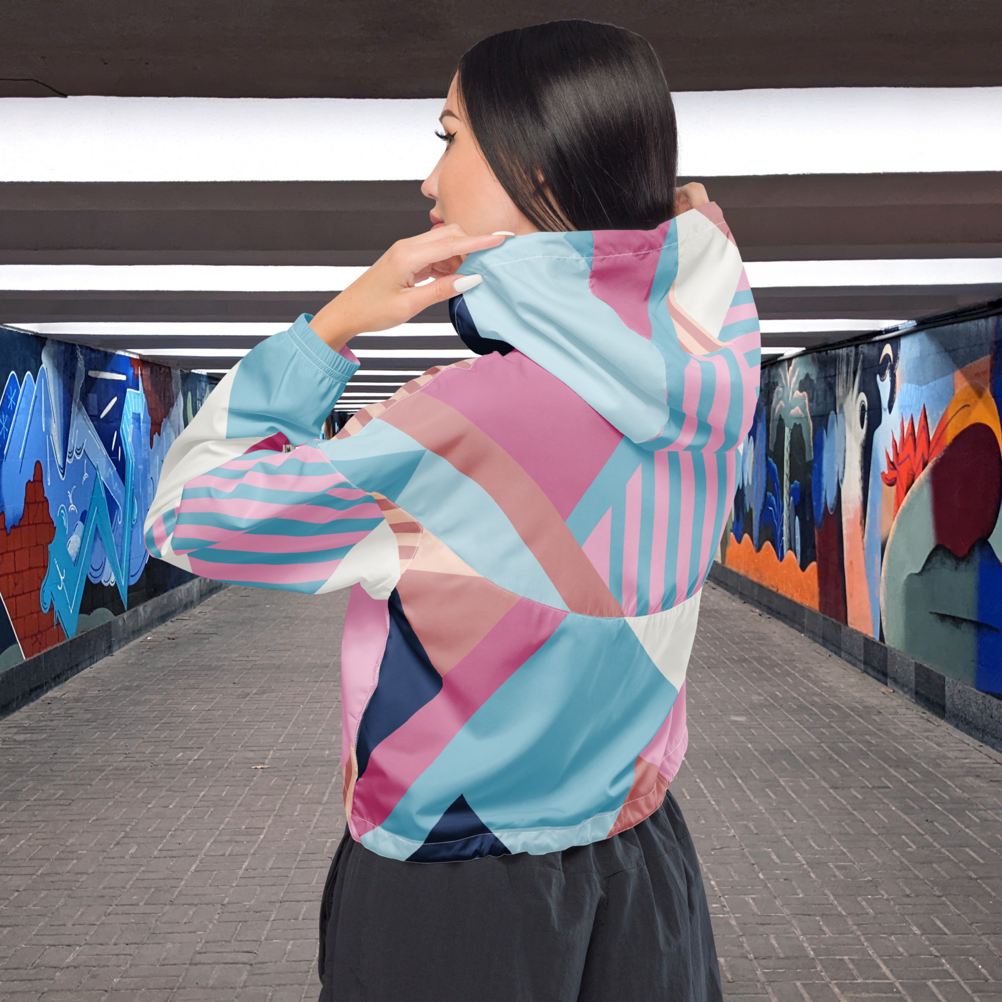 Ein Damen Cropped Windbreaker mit einem lebhaften, geometrischen Muster aus verschiedenen Dreiecken und Rechtecken in Pastellfarben wie Hellblau, Rosa, Beige, Dunkelblau und Weiß. Die Jacke hat eine Kapuze, einen halben Reißverschluss, weiße Kordeln und einen verstellbaren Saum. Mockup Girl Rückseite. By Just a Girl.