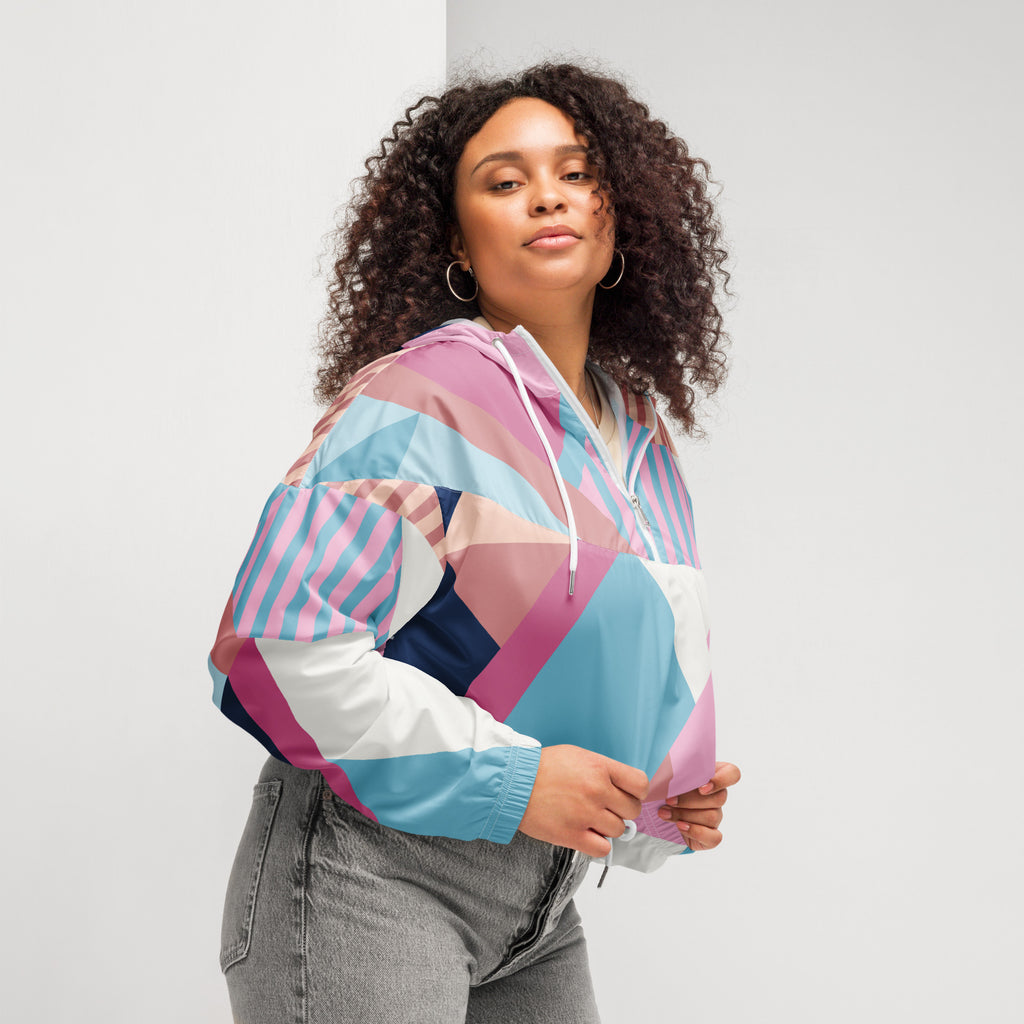 Ein Damen Cropped Windbreaker mit einem lebhaften, geometrischen Muster aus verschiedenen Dreiecken und Rechtecken in Pastellfarben wie Hellblau, Rosa, Beige, Dunkelblau und Weiß. Die Jacke hat eine Kapuze, einen halben Reißverschluss, weiße Kordeln und einen verstellbaren Saum. Mockup woman seiltich rechts.  By Just a Girl.