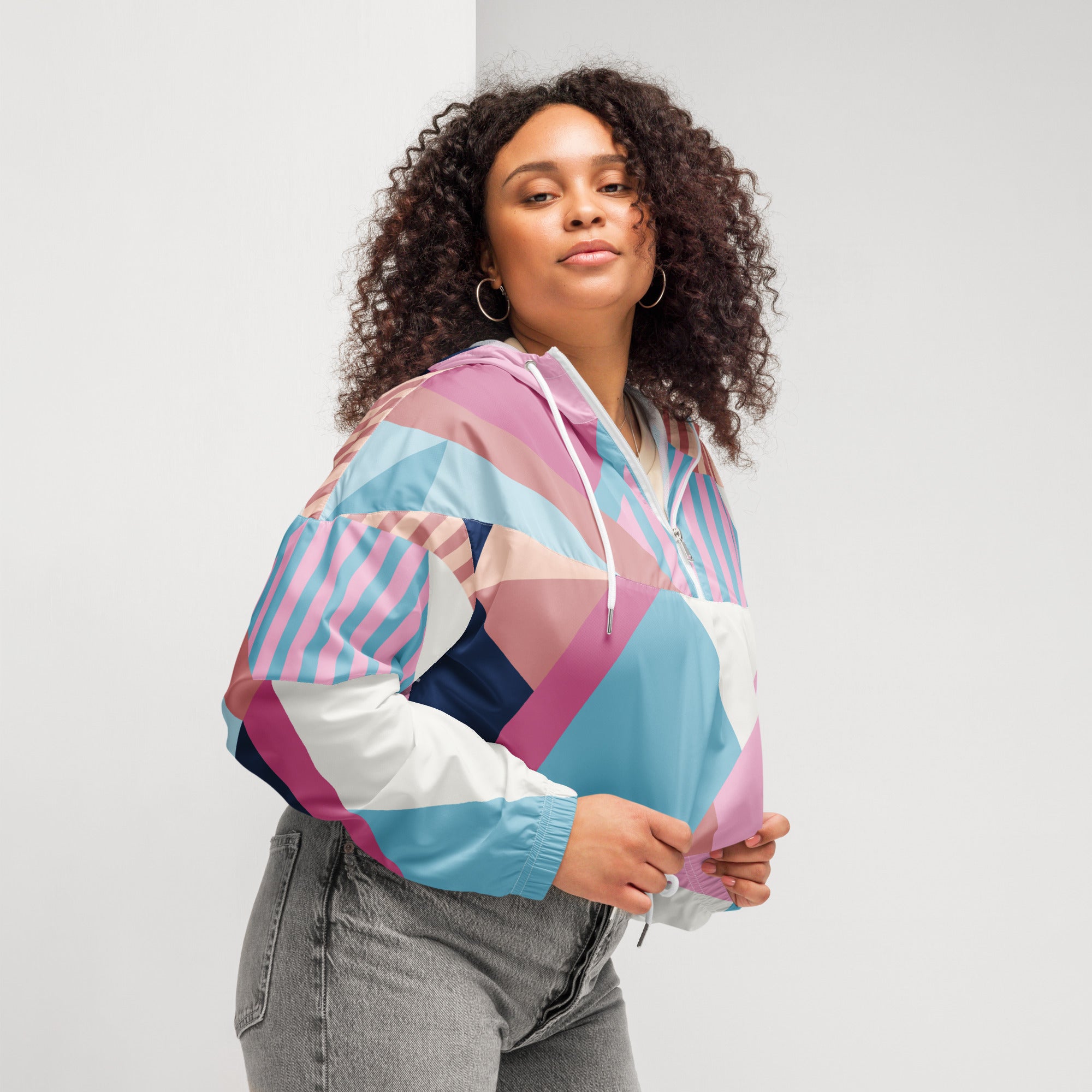 Ein Damen Cropped Windbreaker mit einem lebhaften, geometrischen Muster aus verschiedenen Dreiecken und Rechtecken in Pastellfarben wie Hellblau, Rosa, Beige, Dunkelblau und Weiß. Die Jacke hat eine Kapuze, einen halben Reißverschluss, weiße Kordeln und einen verstellbaren Saum. Mockup woman seiltich rechts.  By Just a Girl.