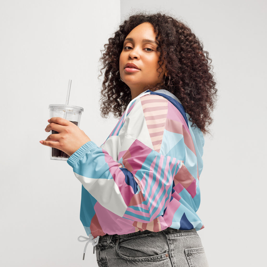 Ein Damen Cropped Windbreaker mit einem lebhaften, geometrischen Muster aus verschiedenen Dreiecken und Rechtecken in Pastellfarben wie Hellblau, Rosa, Beige, Dunkelblau und Weiß. Die Jacke hat eine Kapuze, einen halben Reißverschluss, weiße Kordeln und einen verstellbaren Saum. Mockup Girl seitlich links. By Just a Girl.
