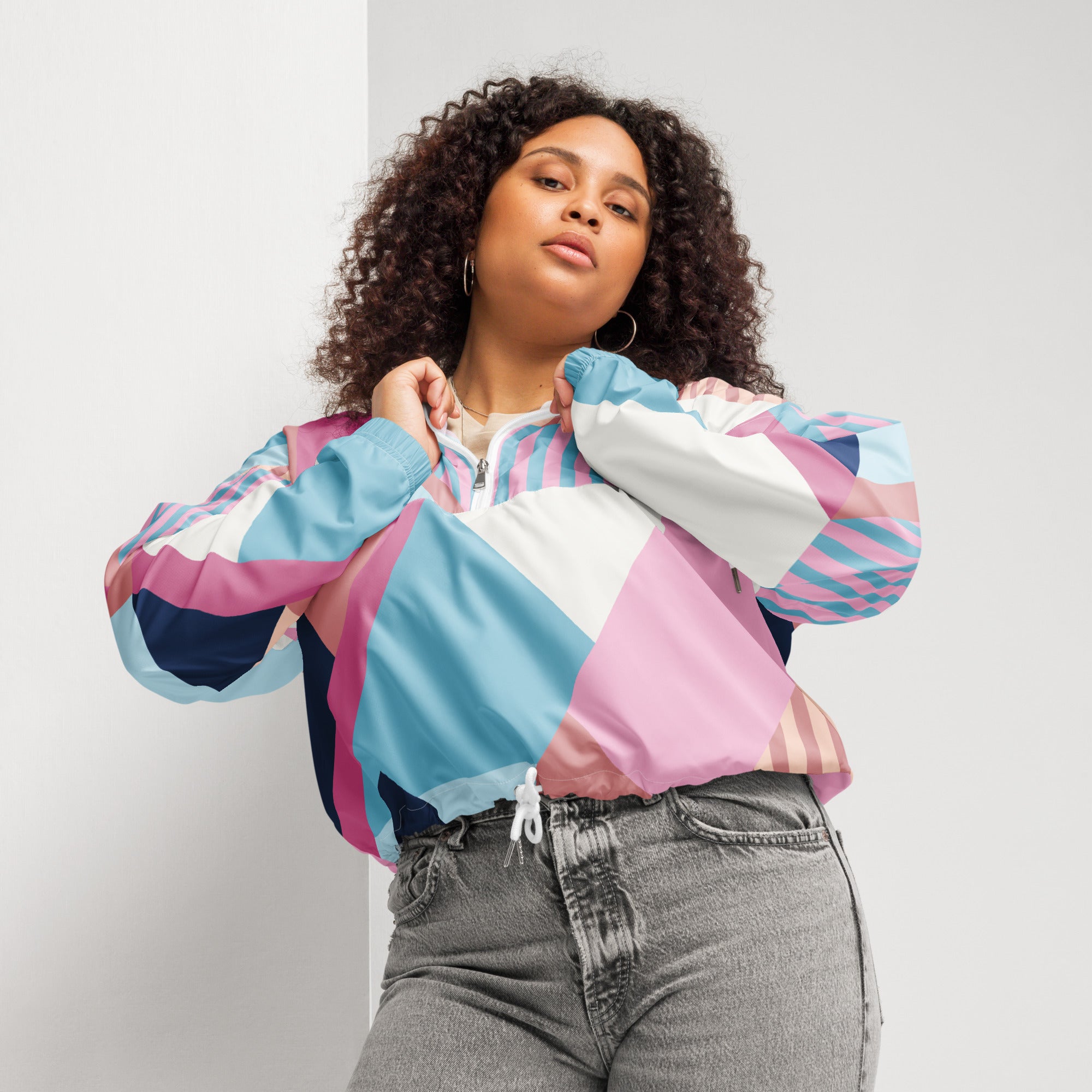 Ein Damen Cropped Windbreaker mit einem lebhaften, geometrischen Muster aus verschiedenen Dreiecken und Rechtecken in Pastellfarben wie Hellblau, Rosa, Beige, Dunkelblau und Weiß. Die Jacke hat eine Kapuze, einen halben Reißverschluss, weiße Kordeln und einen verstellbaren Saum. Mockup woman Vorderseite. By Just a Girl.