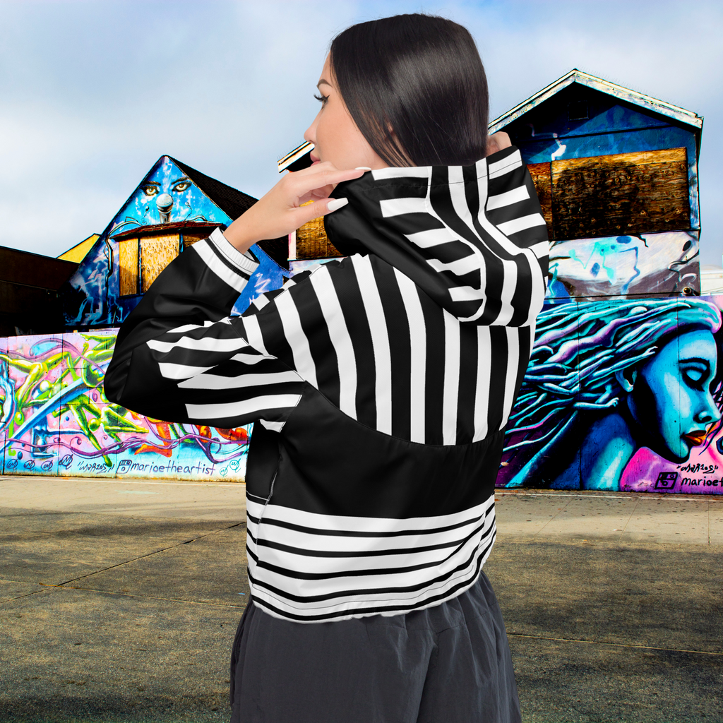 Ein Damen Cropped Windbreaker in Schwarz und Weiß mit einem markanten vertikalen und horizontalen Streifenmuster. Der obere Bereich und die Kapuze zeigen vertikale Streifen, während die Mitte schwarz ist und die untere Hälfte breite horizontale Streifen aufweist. Die Jacke hat einen halben Reißverschluss, schwarze Kordeln und einen verstellbaren Saum. Mockup Girl hintere ansicht By Just a Girl.