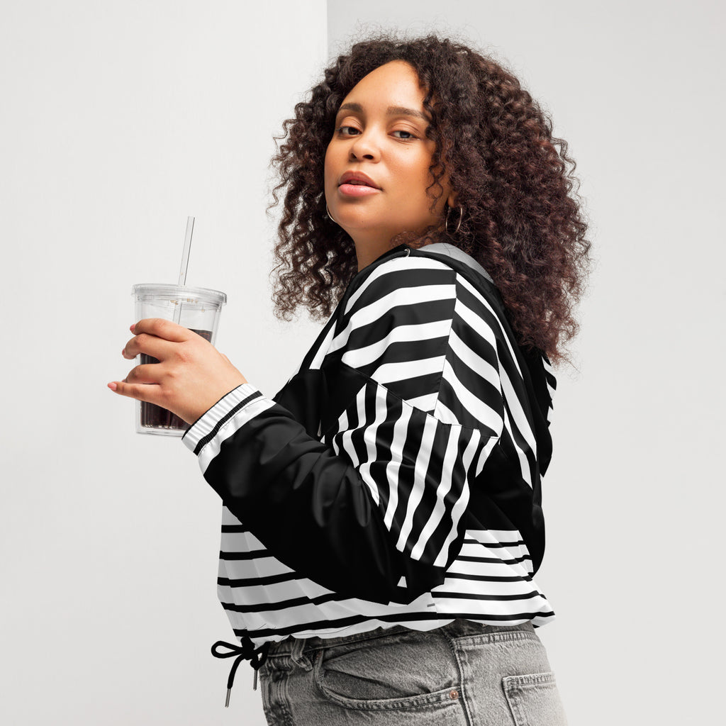 Ein Damen Cropped Windbreaker in Schwarz und Weiß mit einem markanten vertikalen und horizontalen Streifenmuster. Der obere Bereich und die Kapuze zeigen vertikale Streifen, während die Mitte schwarz ist und die untere Hälfte breite horizontale Streifen aufweist. Die Jacke hat einen halben Reißverschluss, schwarze Kordeln und einen verstellbaren Saum. Mockup woman linke seiten ansicht By Just a Girl.