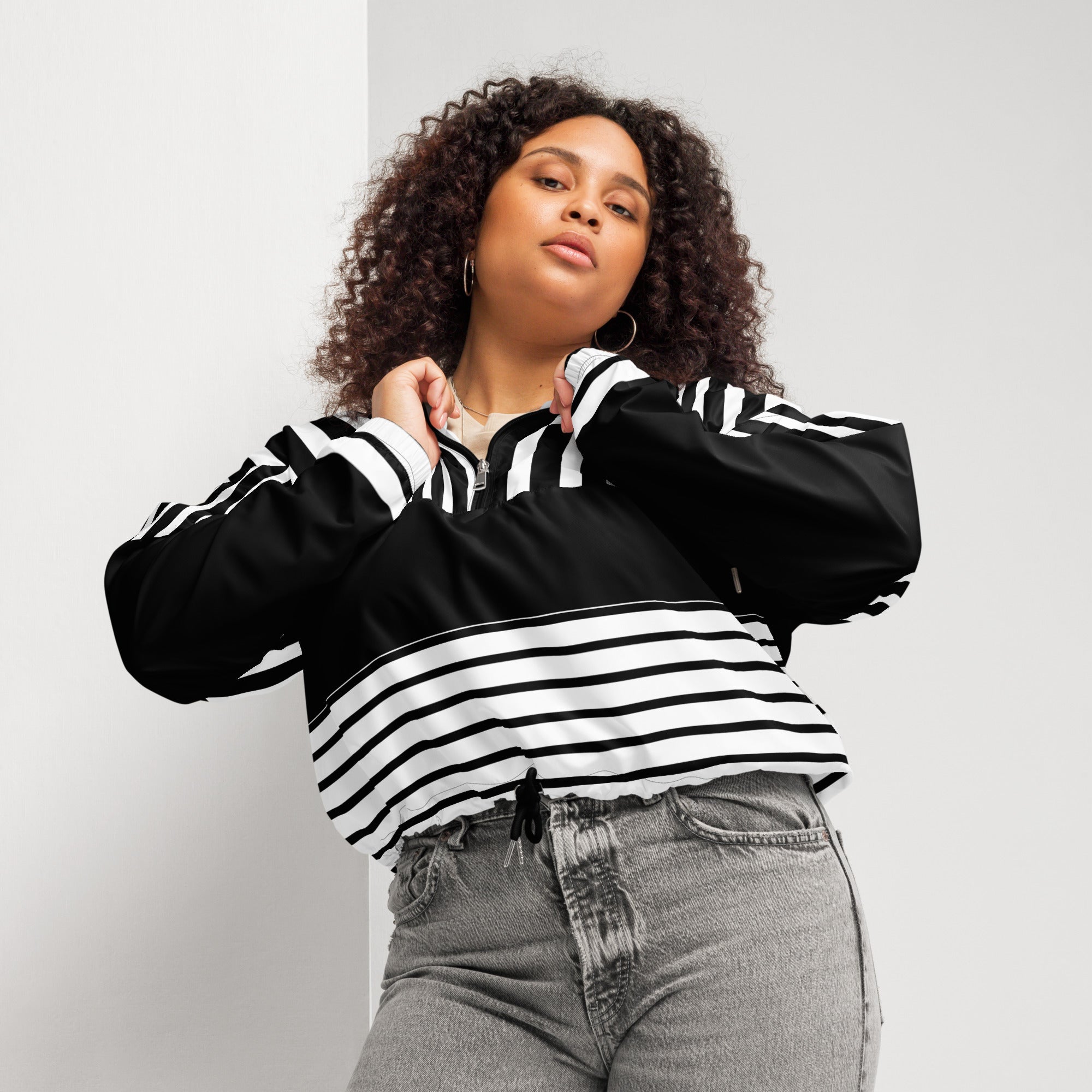 Ein Damen Cropped Windbreaker in Schwarz und Weiß mit einem markanten vertikalen und horizontalen Streifenmuster. Der obere Bereich und die Kapuze zeigen vertikale Streifen, während die Mitte schwarz ist und die untere Hälfte breite horizontale Streifen aufweist. Die Jacke hat einen halben Reißverschluss, schwarze Kordeln und einen verstellbaren Saum. Mockup woman vordere ansicht By Just a Girl.