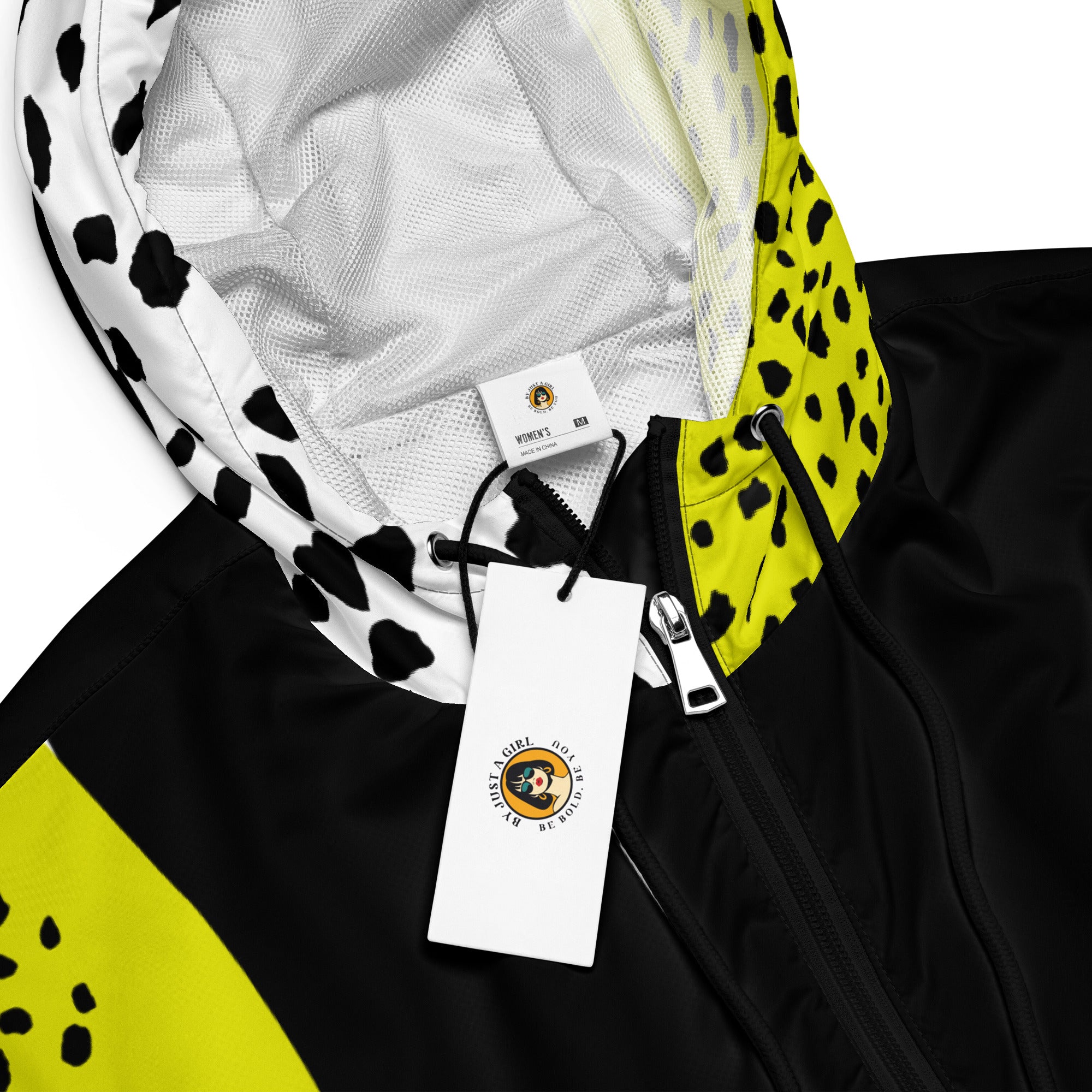 Ein Damen Cropped Windbreaker in Schwarz, Gelb und Weiß mit einem auffälligen, asymmetrischen Muster aus großen gelben Formen mit schwarzen Punkten, schwarzen Segmenten und weißen Bereichen mit schwarzen Punkten. Die Jacke hat eine Kapuze mit gelb-weißem Muster, einen halben Reißverschluss, schwarze Kordeln und einen verstellbaren Saum.  By Just a Girl