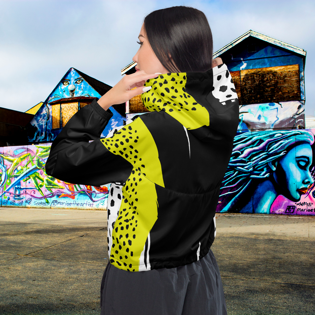 Ein Damen Cropped Windbreaker in Schwarz, Gelb und Weiß mit einem auffälligen, asymmetrischen Muster aus großen gelben Formen mit schwarzen Punkten, schwarzen Segmenten und weißen Bereichen mit schwarzen Punkten. Die Jacke hat eine Kapuze mit gelb-weißem Muster, einen halben Reißverschluss, schwarze Kordeln und einen verstellbaren Saum. Mockup Girl rück ansicht By Just a Girl