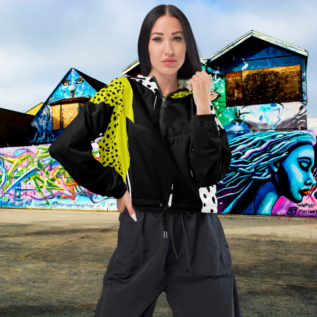 Ein Damen Cropped Windbreaker in Schwarz, Gelb und Weiß mit einem auffälligen, asymmetrischen Muster aus großen gelben Formen mit schwarzen Punkten, schwarzen Segmenten und weißen Bereichen mit schwarzen Punkten. Die Jacke hat eine Kapuze mit gelb-weißem Muster, einen halben Reißverschluss, schwarze Kordeln und einen verstellbaren Saum. Mockup Girl frontansicht By Just a Girl