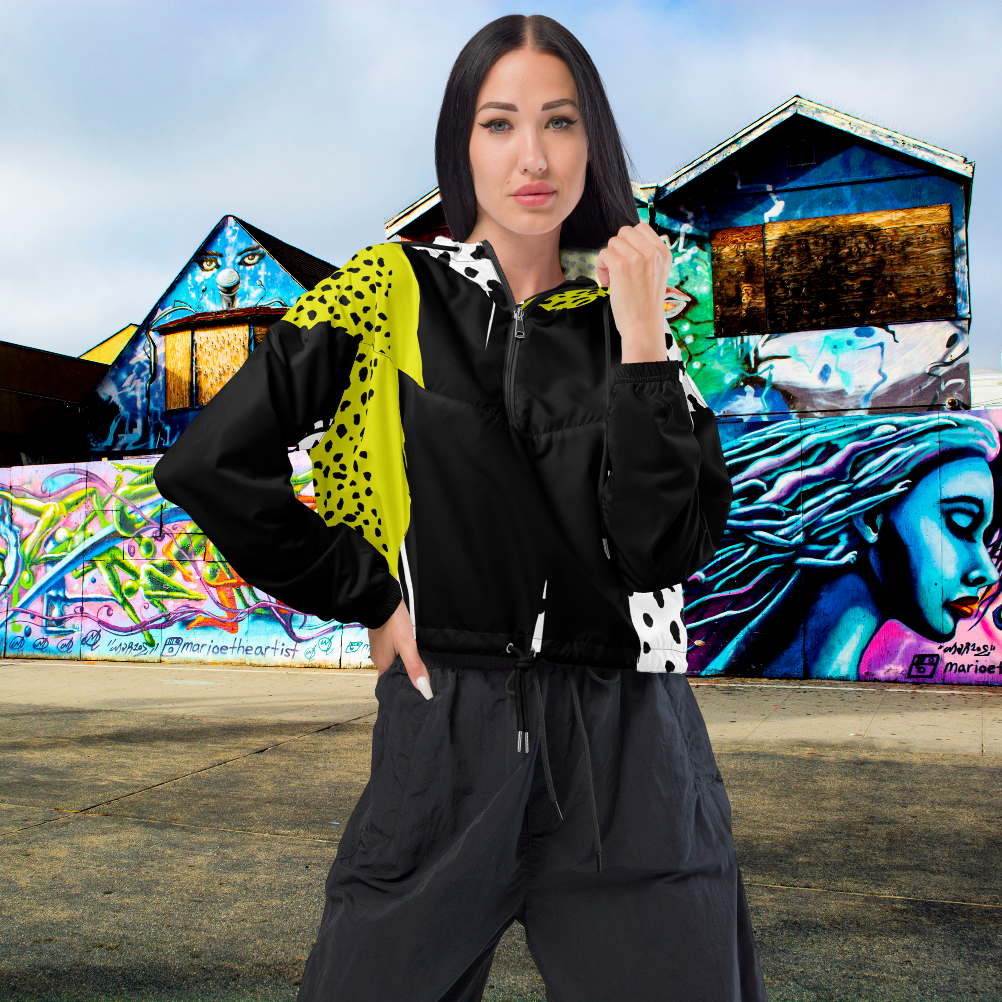 Ein Damen Cropped Windbreaker in Schwarz, Gelb und Weiß mit einem auffälligen, asymmetrischen Muster aus großen gelben Formen mit schwarzen Punkten, schwarzen Segmenten und weißen Bereichen mit schwarzen Punkten. Die Jacke hat eine Kapuze mit gelb-weißem Muster, einen halben Reißverschluss, schwarze Kordeln und einen verstellbaren Saum. Mockup Girl frontansicht By Just a Girl