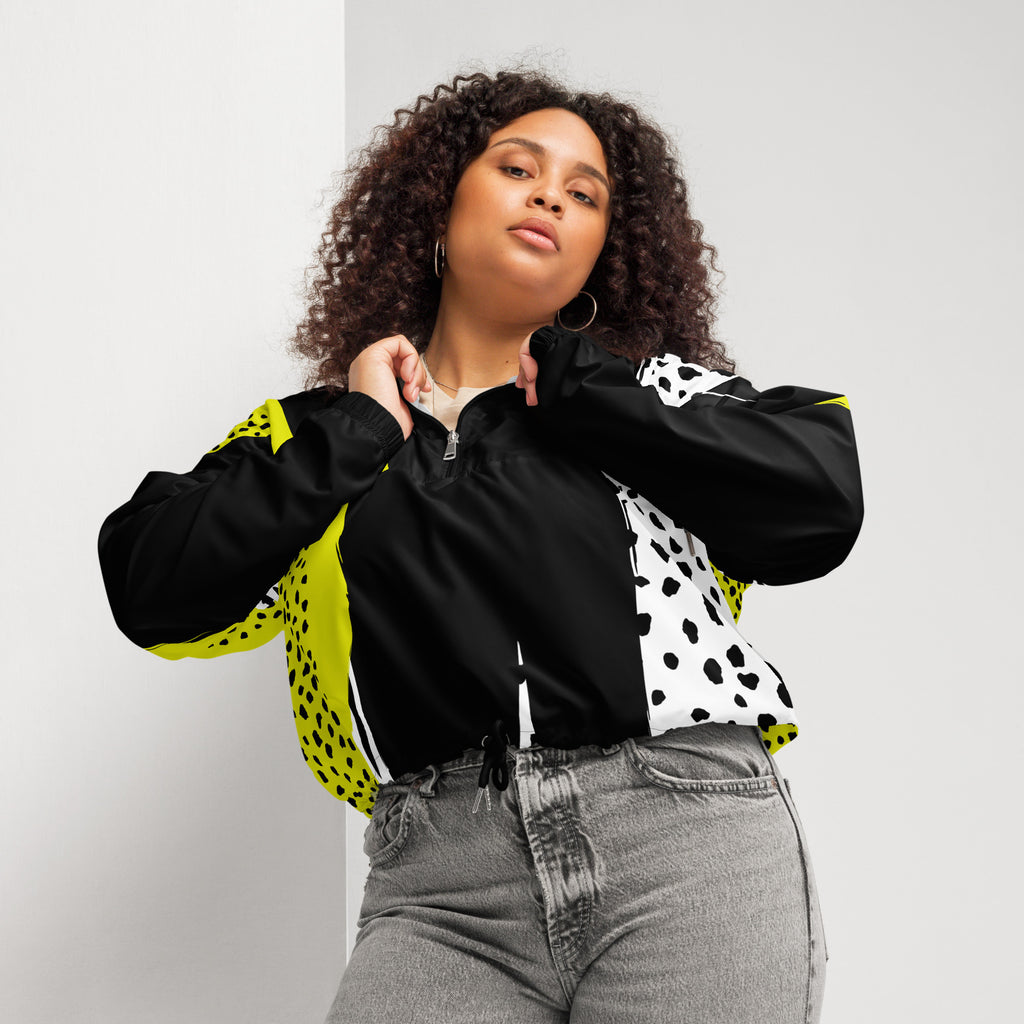 Ein Damen Cropped Windbreaker in Schwarz, Gelb und Weiß mit einem auffälligen, asymmetrischen Muster aus großen gelben Formen mit schwarzen Punkten, schwarzen Segmenten und weißen Bereichen mit schwarzen Punkten. Die Jacke hat eine Kapuze mit gelb-weißem Muster, einen halben Reißverschluss, schwarze Kordeln und einen verstellbaren Saum. Mockup woman frontansicht By Just a Girl