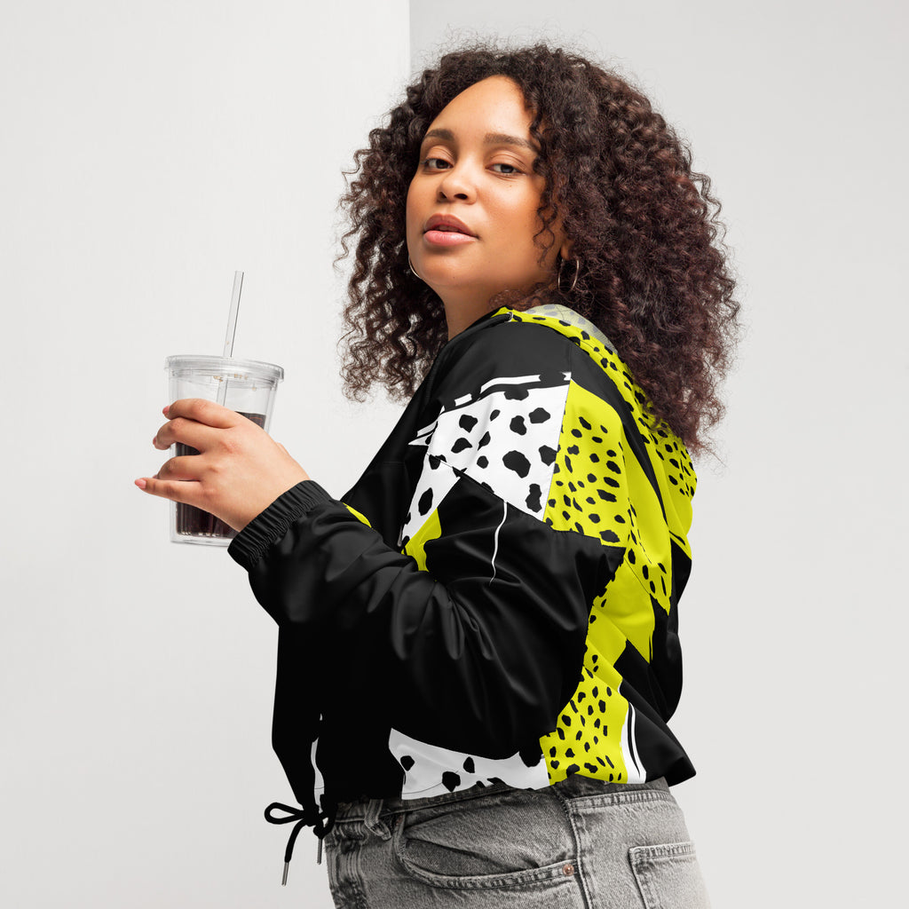 Ein Damen Cropped Windbreaker in Schwarz, Gelb und Weiß mit einem auffälligen, asymmetrischen Muster aus großen gelben Formen mit schwarzen Punkten, schwarzen Segmenten und weißen Bereichen mit schwarzen Punkten. Die Jacke hat eine Kapuze mit gelb-weißem Muster, einen halben Reißverschluss, schwarze Kordeln und einen verstellbaren Saum. Mockup woman linke seiten ansicht By Just a Girl