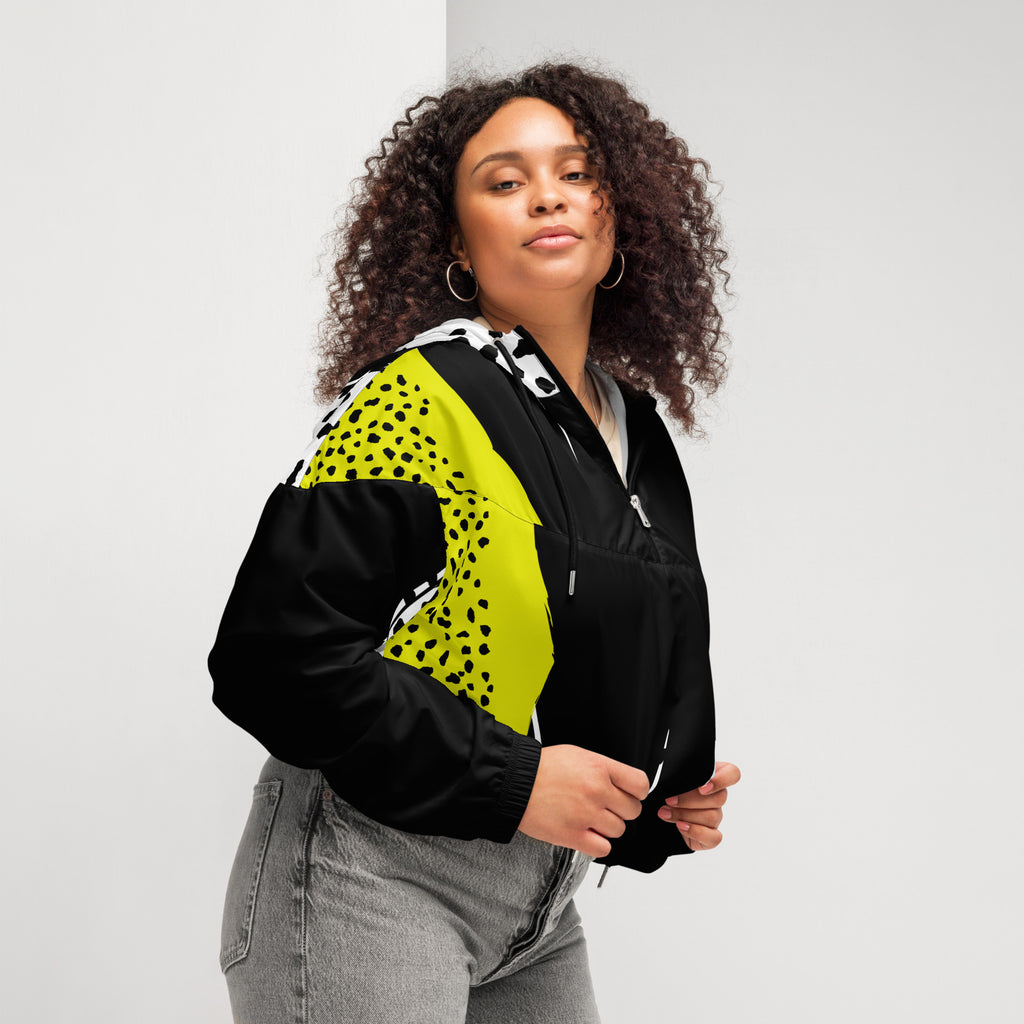 Ein Damen Cropped Windbreaker in Schwarz, Gelb und Weiß mit einem auffälligen, asymmetrischen Muster aus großen gelben Formen mit schwarzen Punkten, schwarzen Segmenten und weißen Bereichen mit schwarzen Punkten. Die Jacke hat eine Kapuze mit gelb-weißem Muster, einen halben Reißverschluss, schwarze Kordeln und einen verstellbaren Saum. Mockup Girl seitliche ansicht rechts By Just a Girl