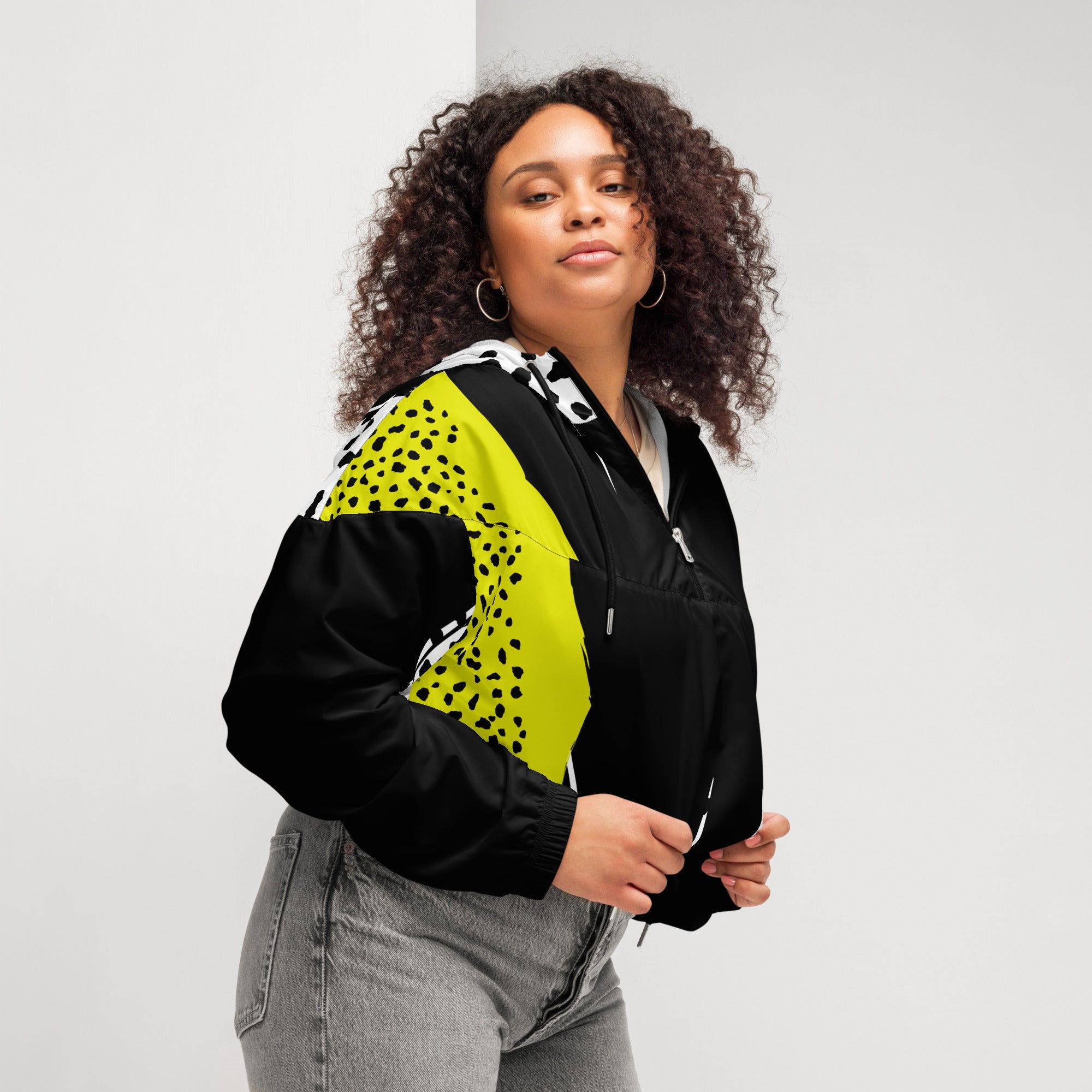 Ein Damen Cropped Windbreaker in Schwarz, Gelb und Weiß mit einem auffälligen, asymmetrischen Muster aus großen gelben Formen mit schwarzen Punkten, schwarzen Segmenten und weißen Bereichen mit schwarzen Punkten. Die Jacke hat eine Kapuze mit gelb-weißem Muster, einen halben Reißverschluss, schwarze Kordeln und einen verstellbaren Saum. Mockup Girl seitliche ansicht rechts By Just a Girl