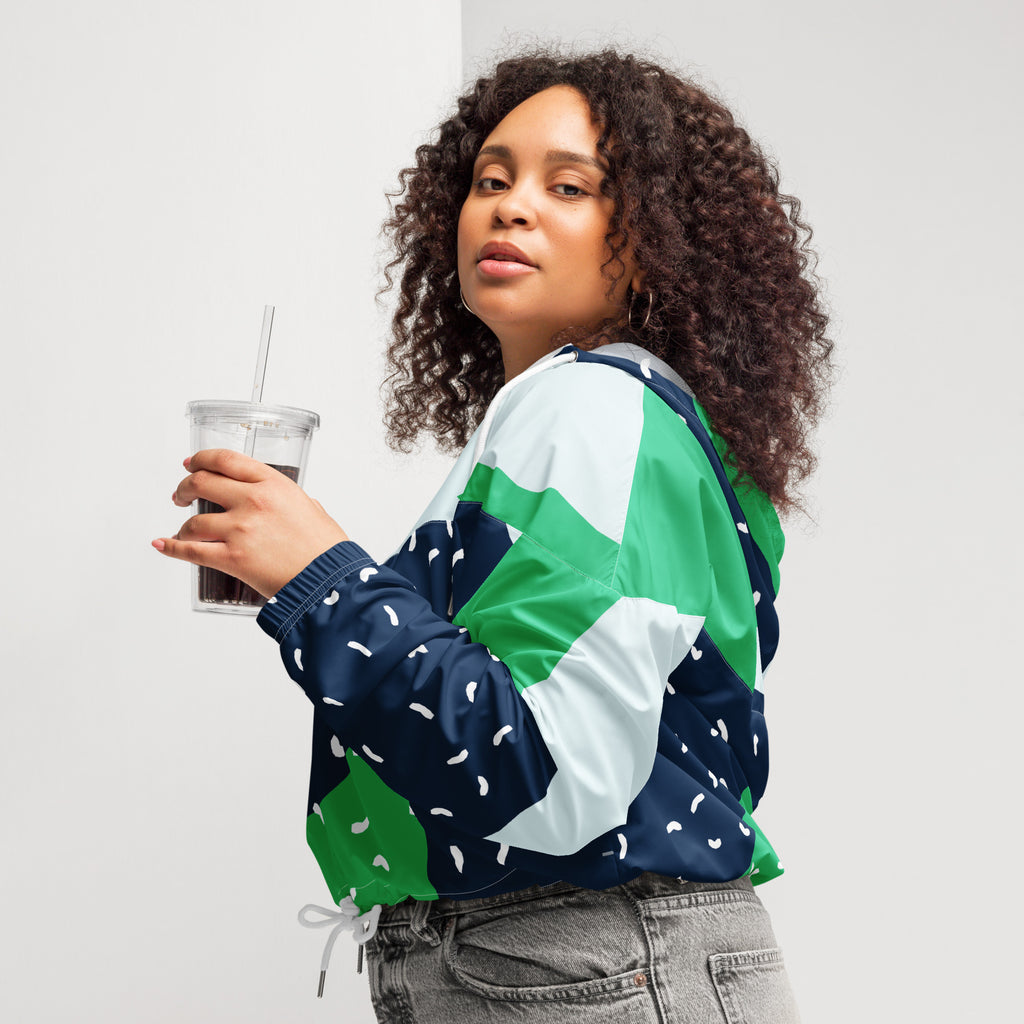 Ein Damen Cropped Windbreaker mit einem modernen, geometrischen Blockmuster in Dunkelblau, hellem Mintgrün und strahlendem Grün. Der dunkelblaue Bereich ist zusätzlich mit kleinen weißen, unregelmäßigen Strichformen versehen. Die Jacke hat eine Kapuze, einen halben Reißverschluss mit weißen Kordeln und einen verstellbaren Saum. Mockup woman seitliche Ansicht links. By Just a Girl.